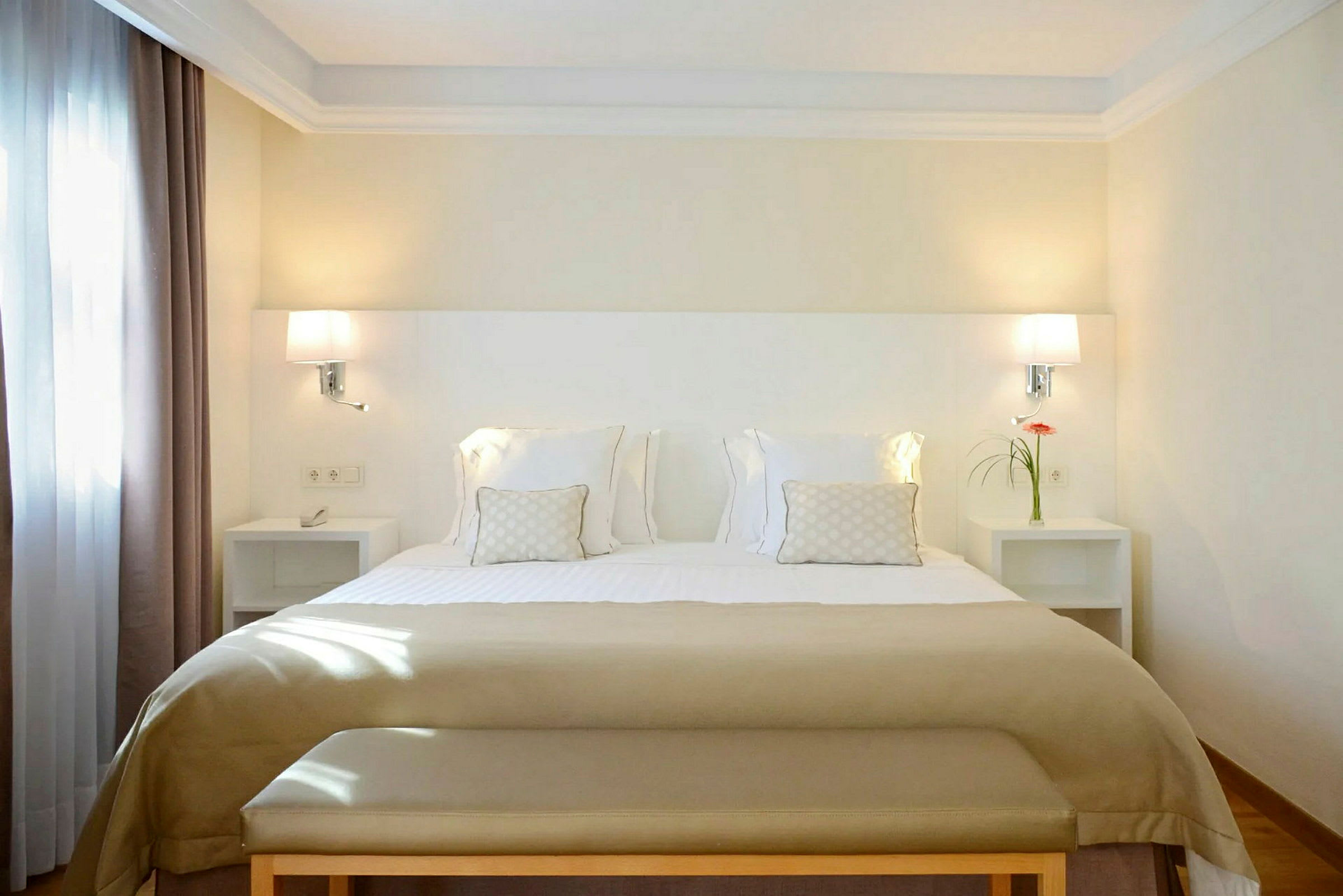 Hotellihuone, jossa on leveä sänky, koristeltu valkoisilla ja beigeillä vuodevaatteilla, sekä yöpöydät lampuilla ja koristelulla. (Tekoälyn luoma teksti)
