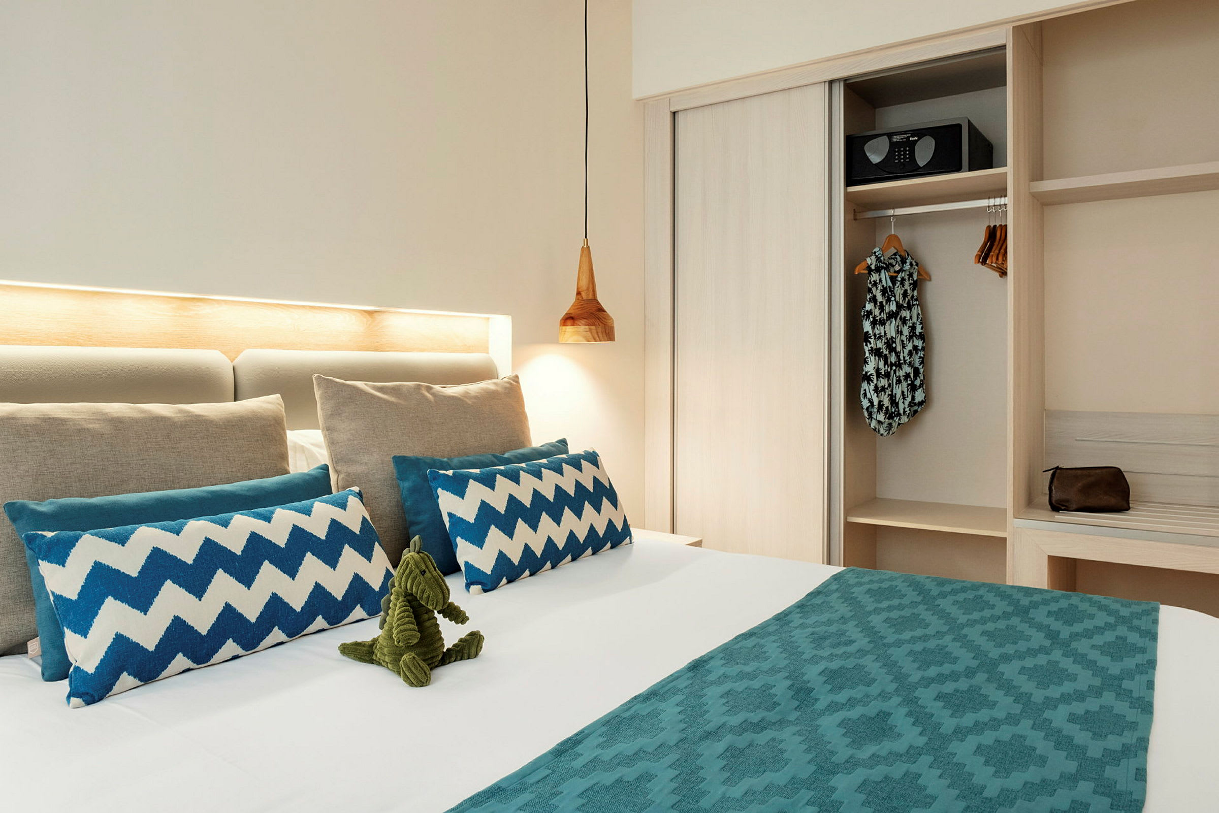 Hotellihuone, jossa on pedattu sänky koristeltuna sinisillä tyynyillä ja vihreällä lelulla. Vieressä näkyy vaatekaappi, jossa roikkuu vaatteita sekä tallelokero. (Tekoälyn luoma teksti)