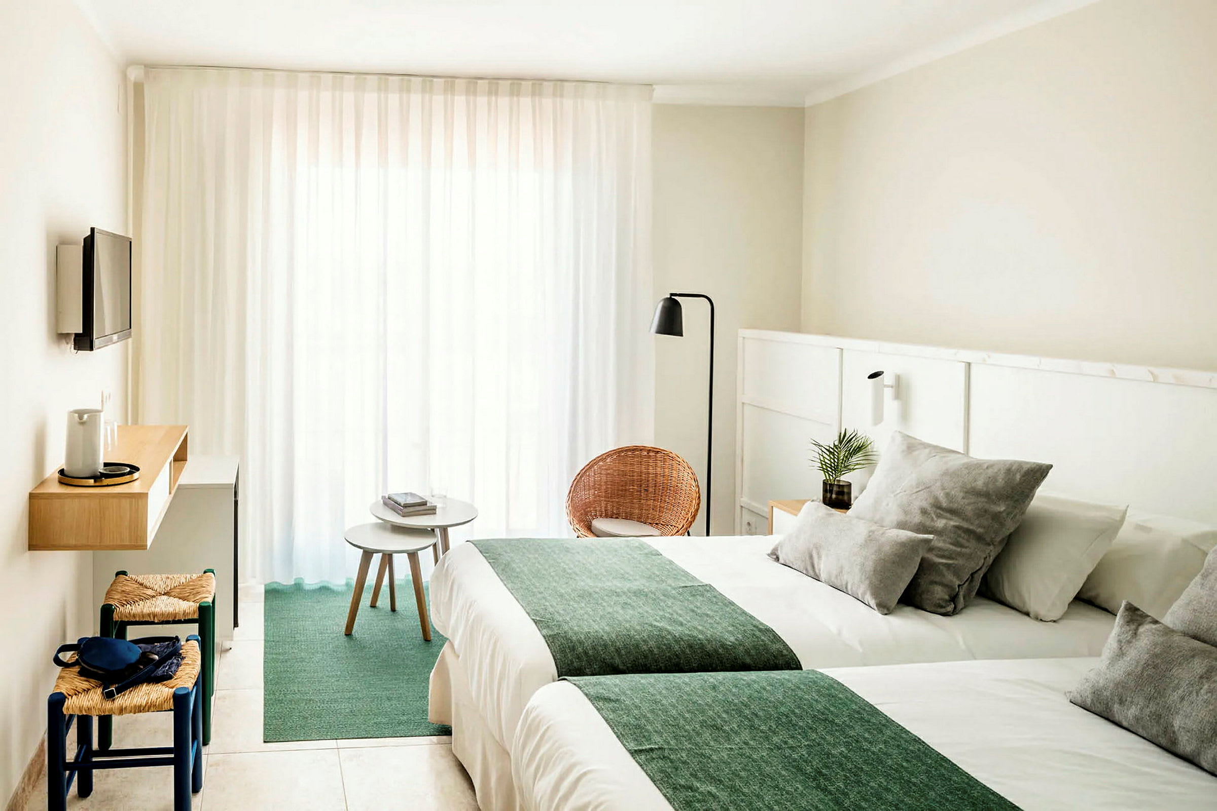 Hotellihuone, jossa on vaaleat seinät, kaksi sänkyä vihreillä peitteillä, pieni istuinryhmä tuoleilla ja pöydällä sekä seinälle kiinnitetty TV. (Tekoälyn luoma teksti)