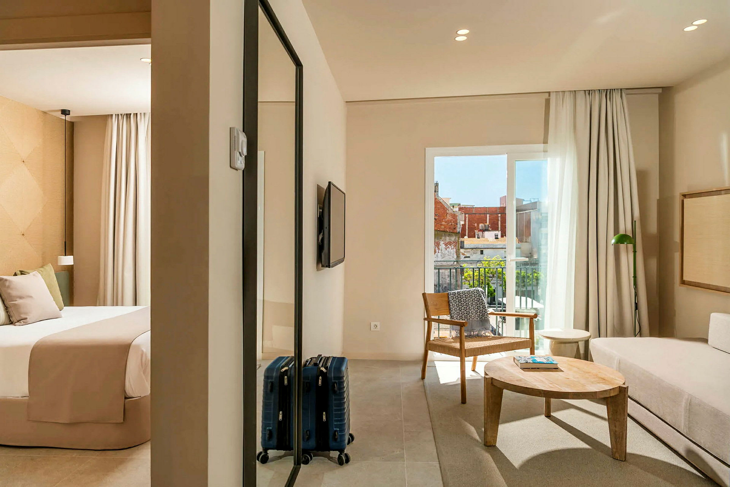 Hotellihuone, jossa on makuuhuone ja olohuone. Makuuhuoneessa on beigeillä vuodevaatteilla varustettu sänky, ja olohuoneessa on sohva, puupöytä ja nojatuoli. Parveke, jolta on näkymä. (Tekoälyn luoma teksti)