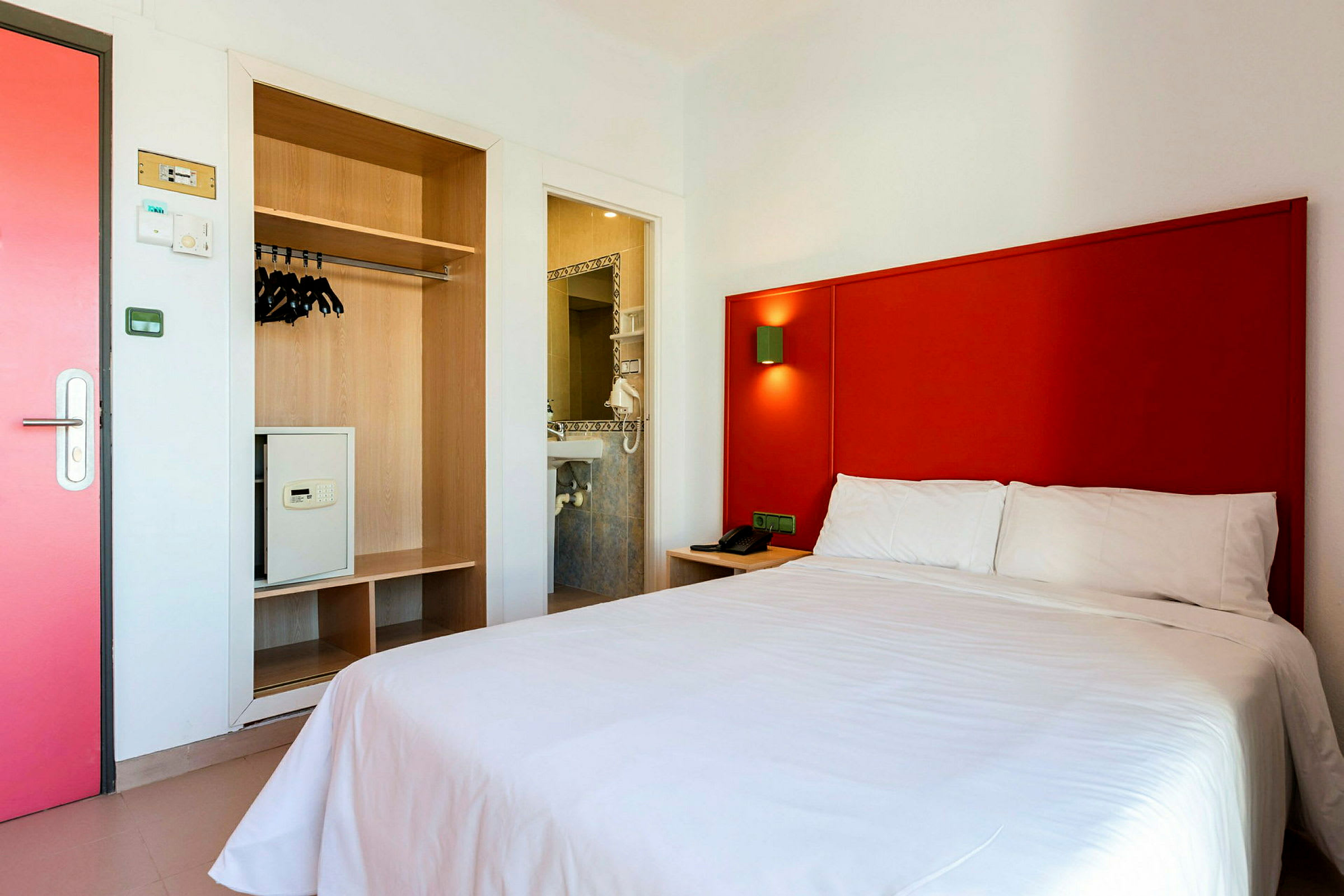 Hotellihuone, jossa punainen sängynpääty, valkoinen sänky ja vaatekaappi tallelokerolla. (Tekoälyn luoma teksti)
