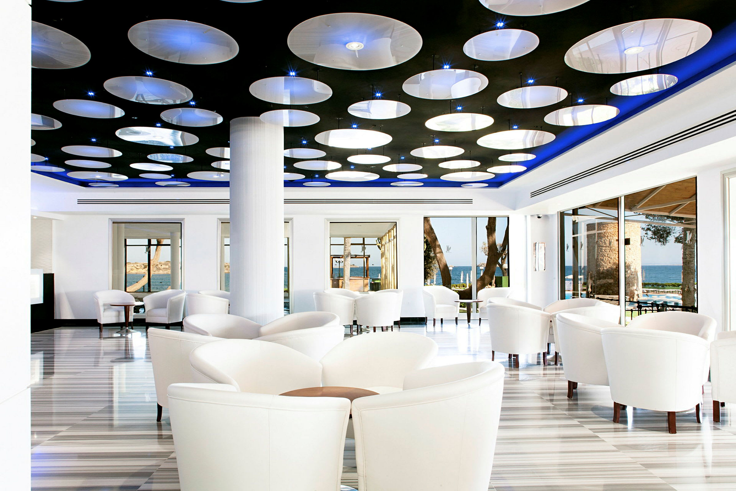 Modernisti sisustettu kahvila tai lounge, jossa on valkoiset tuolit, suuret ikkunat ja pyöreistä valoista koostuva kattodesign. (Tekoälyn luoma teksti)