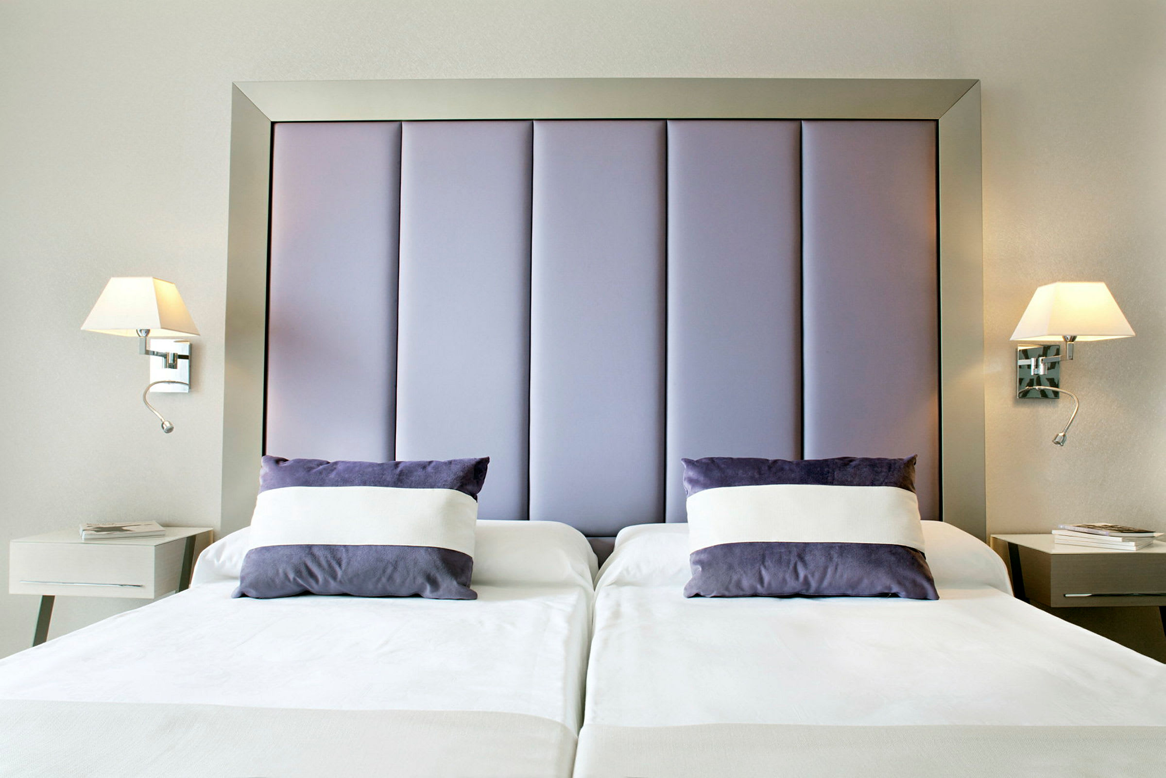 Hotellihuone, jossa kaksi yhden hengen vuodetta, joissa on valkoiset lakanat ja violetit tyynyt. Seinälamput ovat vuoteiden molemmilla puolilla, ja jokaisella vuoteella on yöpöytä. (Tekoälyn luoma teksti)