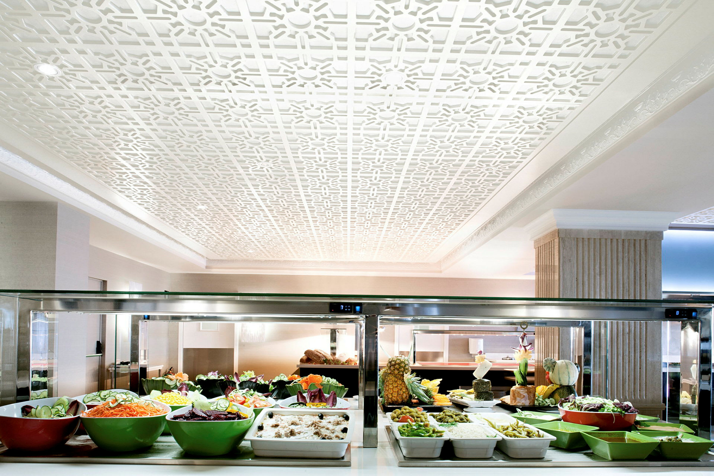 Buffet, jossa on erilaisia salaatteja ja ruokia kulhoissa ja astioissa, ympäröitynä elegantisti sisustetulla huoneella, jossa on valkoinen koristelu katossa. (Tekoälyn luoma teksti)