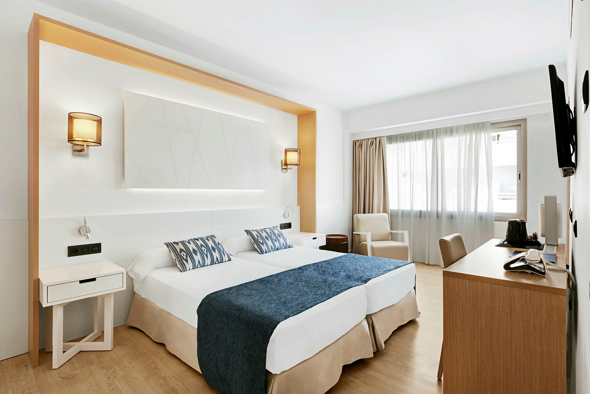Hotellihuone, jossa kaksi sänkyä sinisillä päiväpeitteillä ja kuvioiduilla tyynyillä, työpöytä, tuoli sekä lepotuoli ikkunan vieressä. (Tekoälyn luoma teksti)