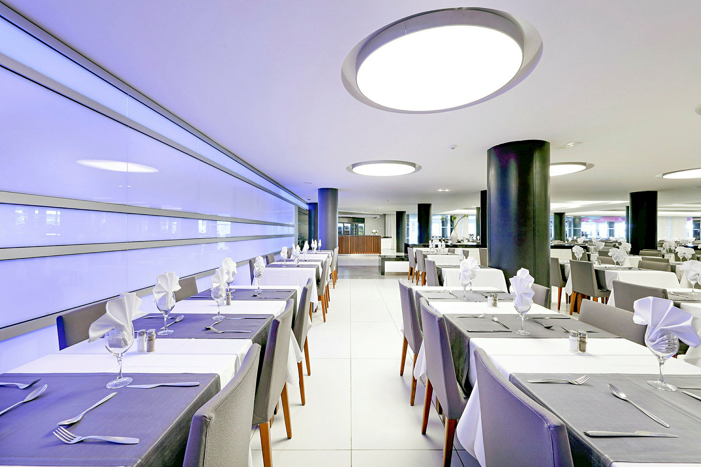 Moderni ravintola katetuilla pöydillä ja valkoisilla tuoleilla. Sisustus sisältää moderneja valaisimia ja valoisan ilmapiirin. (Tekoälyn luoma teksti)