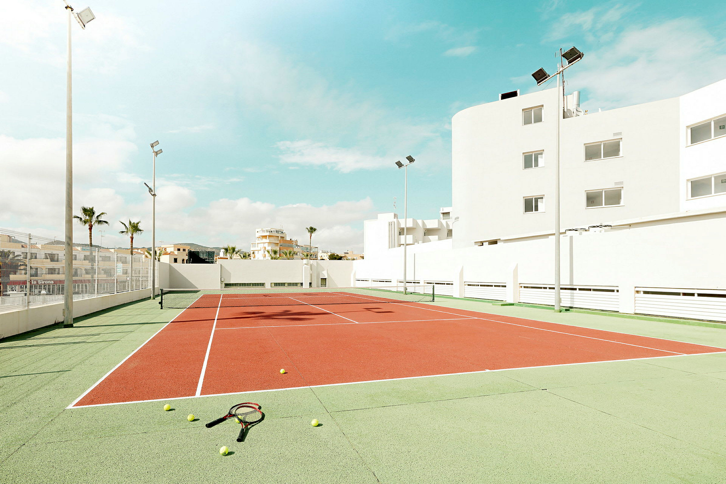 Tenniskenttä, jota ympäröivät rakennukset aurinkoisen taivaan alla, ja kentällä maassa tennispalloja ja mailoja. (Tekoälyn luoma teksti)