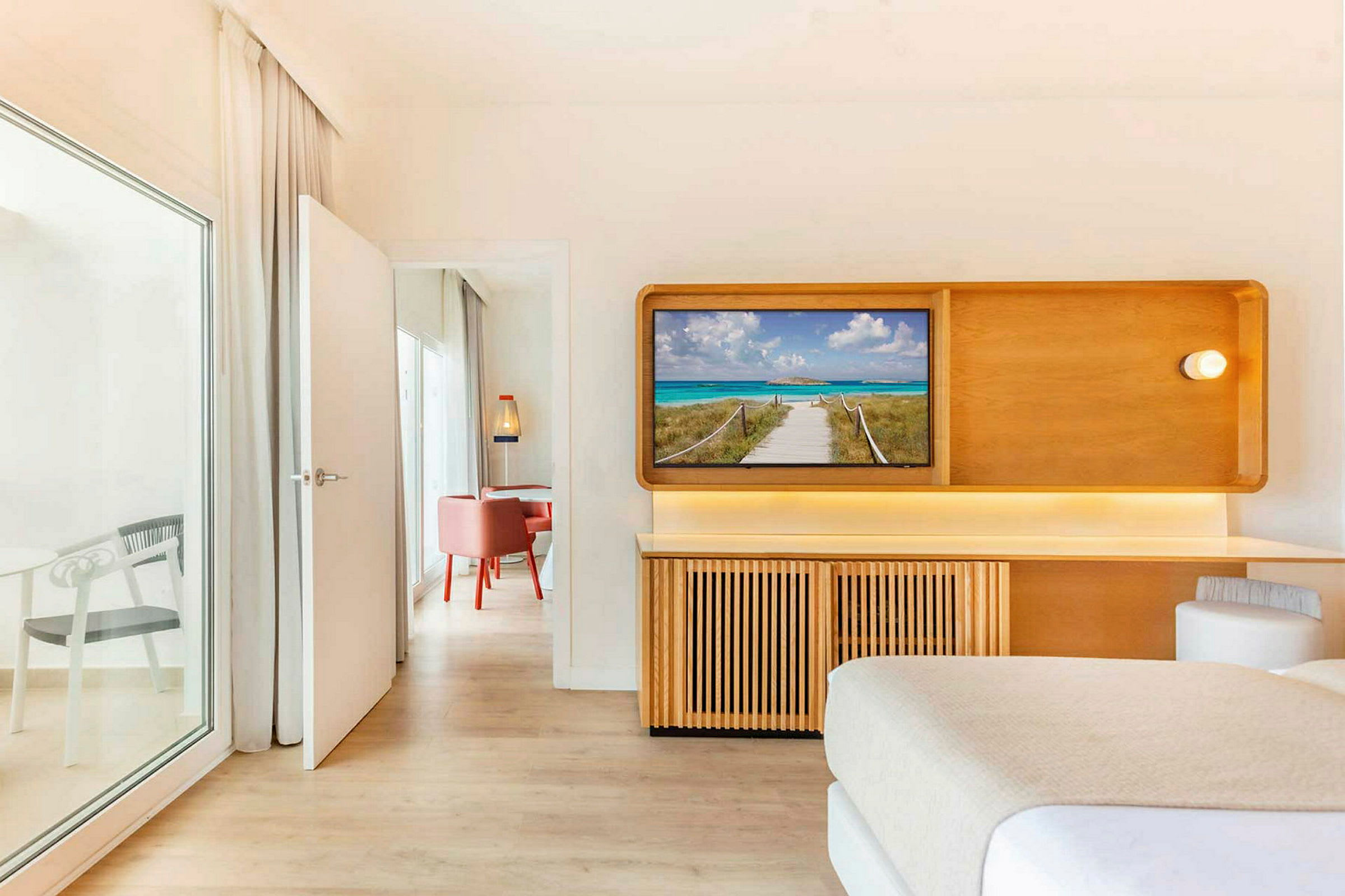 Hotellihuone, jossa on televisio, joka näyttää rantalaiturin. Ovi johtaa viereiseen huoneeseen, jossa on punaisia tuoleja. (Tekoälyn luoma teksti)