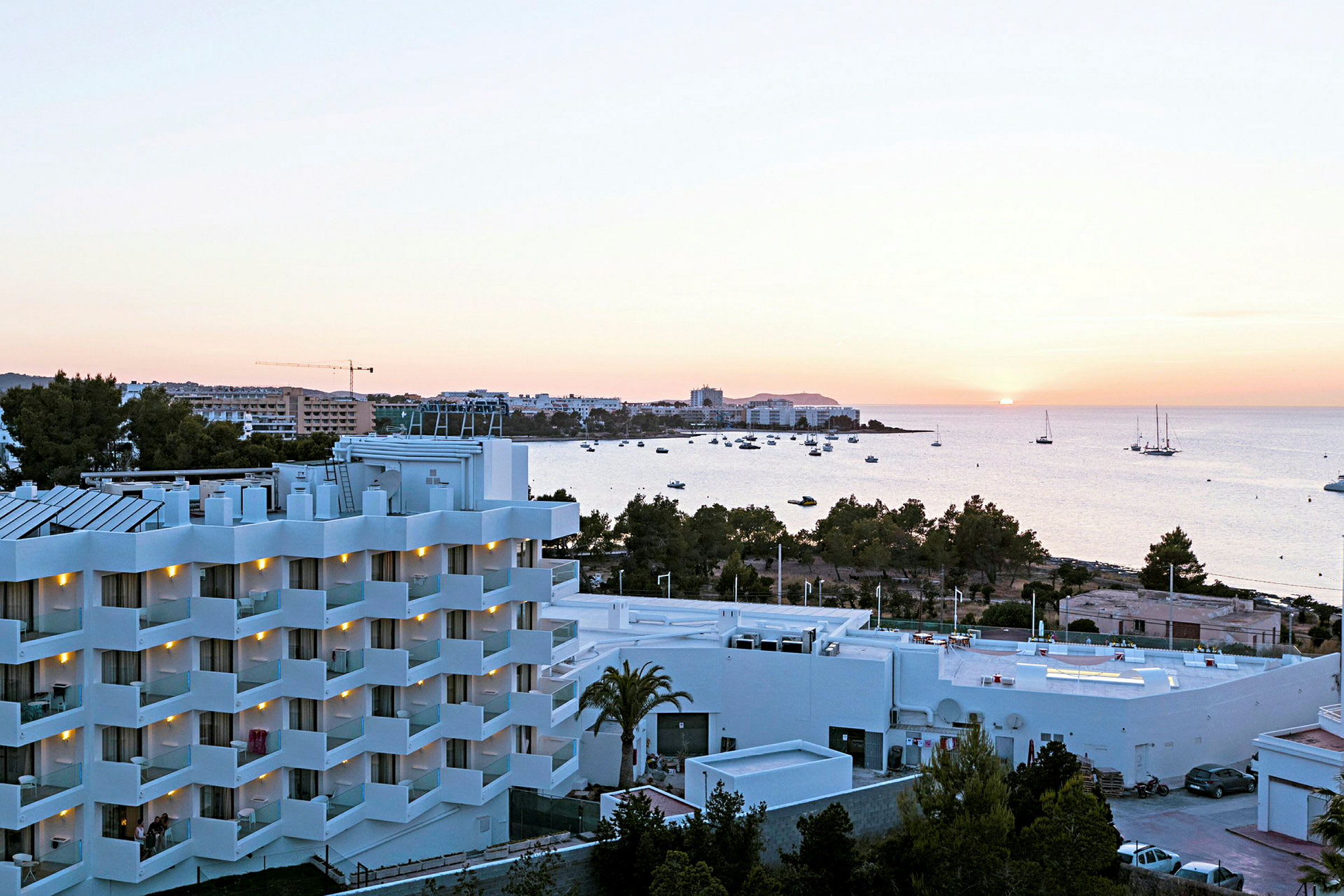 Näkymä hotellista, jossa on valkoiset julkisivut ja parvekkeet, vehreyden ympäröimänä, taustalla meri ja auringonlasku. (Tekoälyn luoma teksti)
