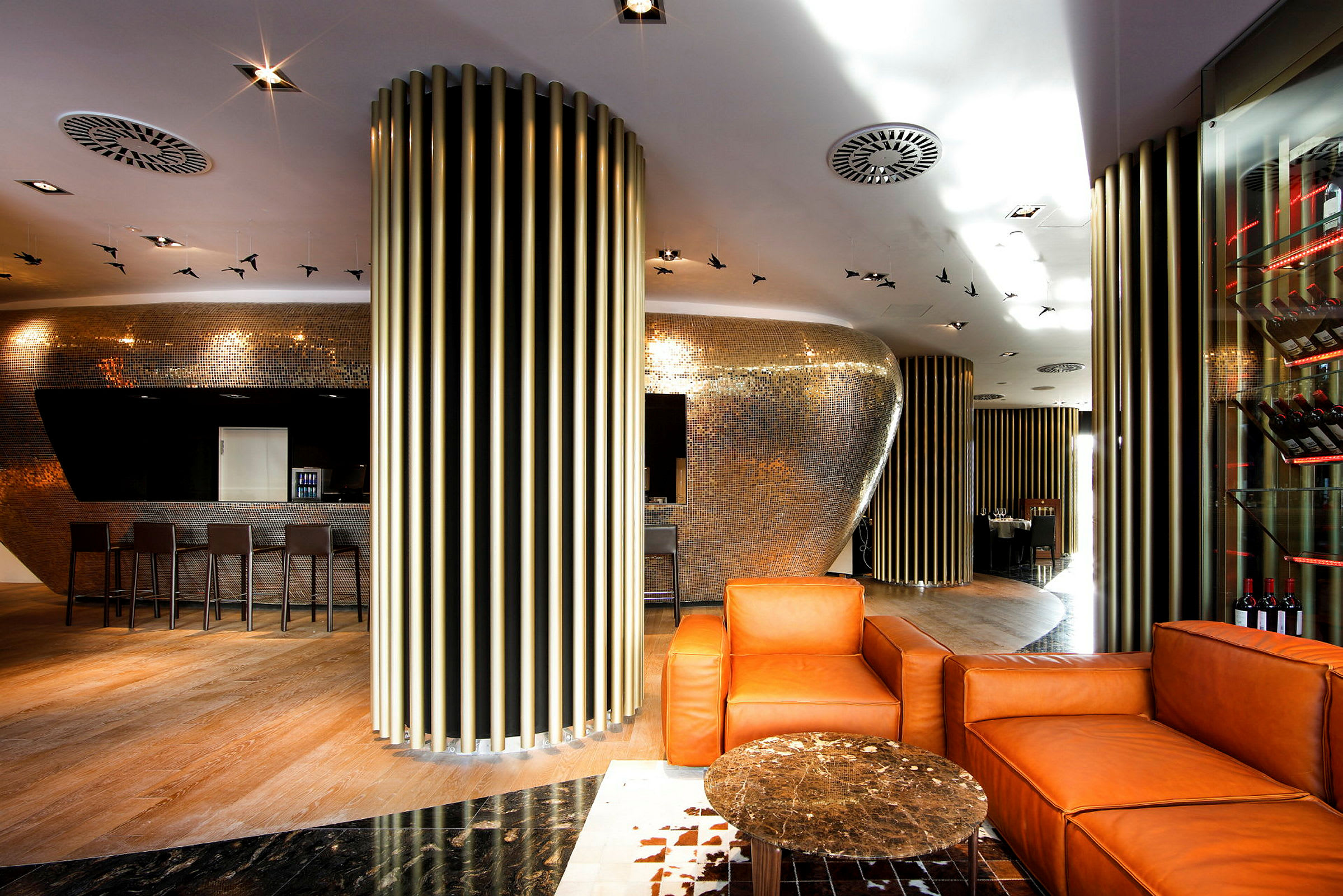 Modernisti sisustettu lounge, jossa oransseja nojatuoleja, pieni pöytä sekä metallikoristeita seinillä ja katossa. (Tekoälyn luoma teksti)