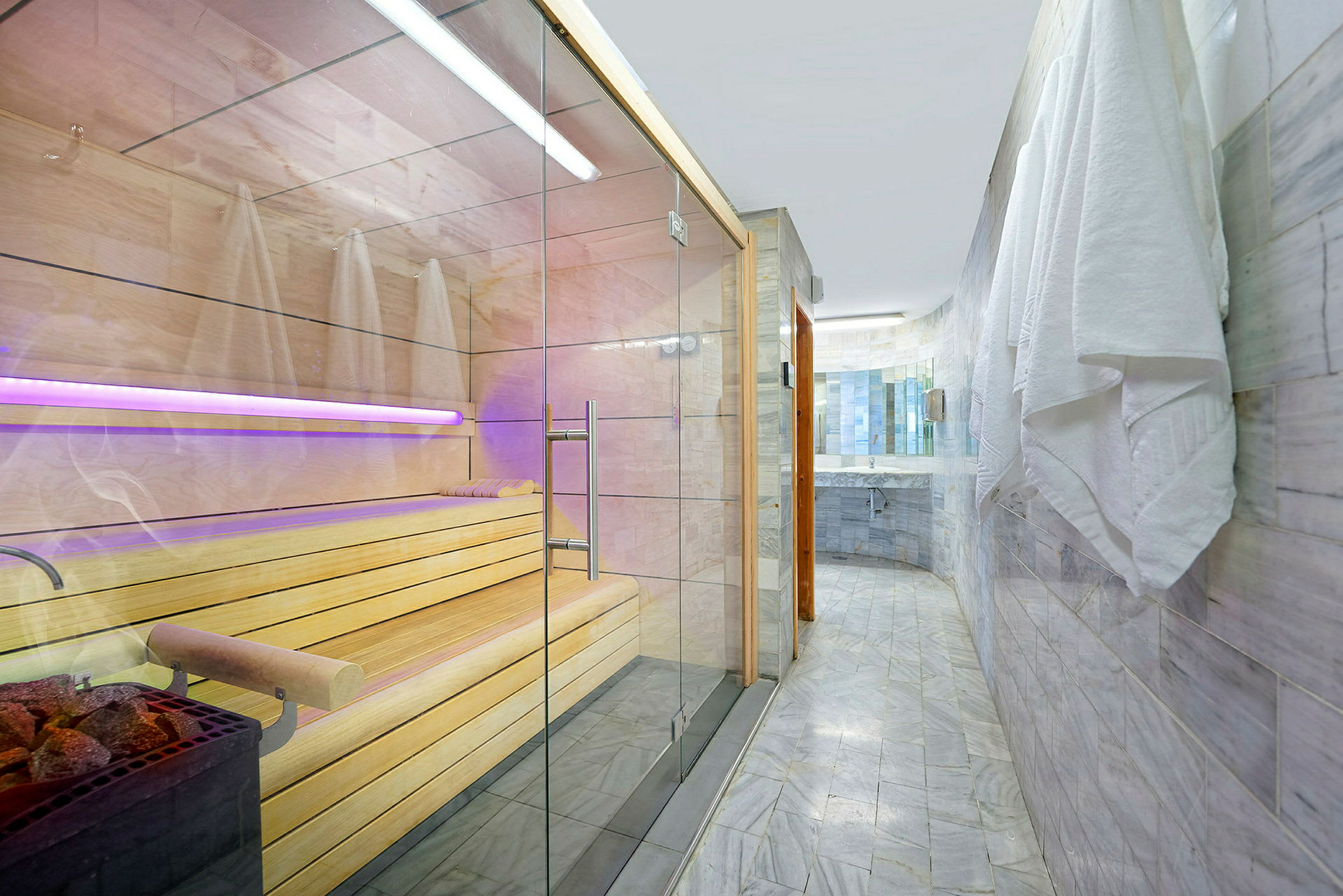 Kuvassa on moderni sauna lasiovilla ja puusisustuksella sekä kylpyhuone laatoitetuilla seinillä ja seinällä roikkuvilla pyyhkeillä. (Tekoälyn luoma teksti)