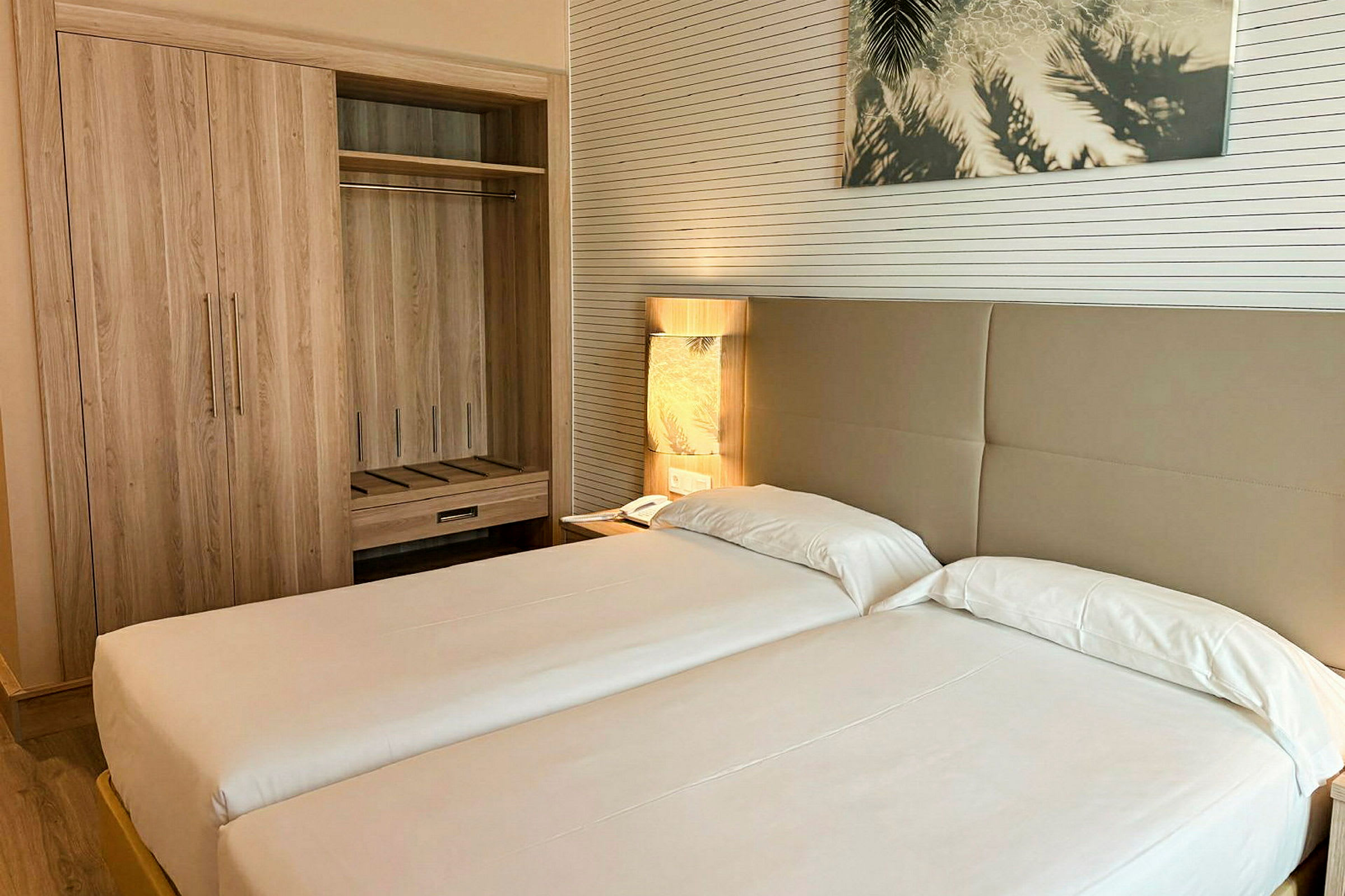 Hotellihuone, jossa kaksi yhden hengen vuodetta, vaatekaappi ja yöpöydällä pöytälamppu. (Tekoälyn luoma teksti)