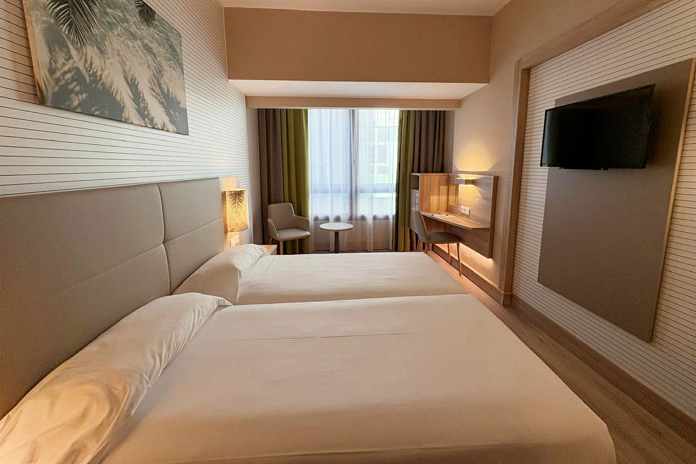 Hotellihuone, jossa kaksi erillistä vuodetta, ikkuna verhoineen, istuinalue pöytineen ja seinään asennettu taulu-tv. (Tekoälyn luoma teksti)