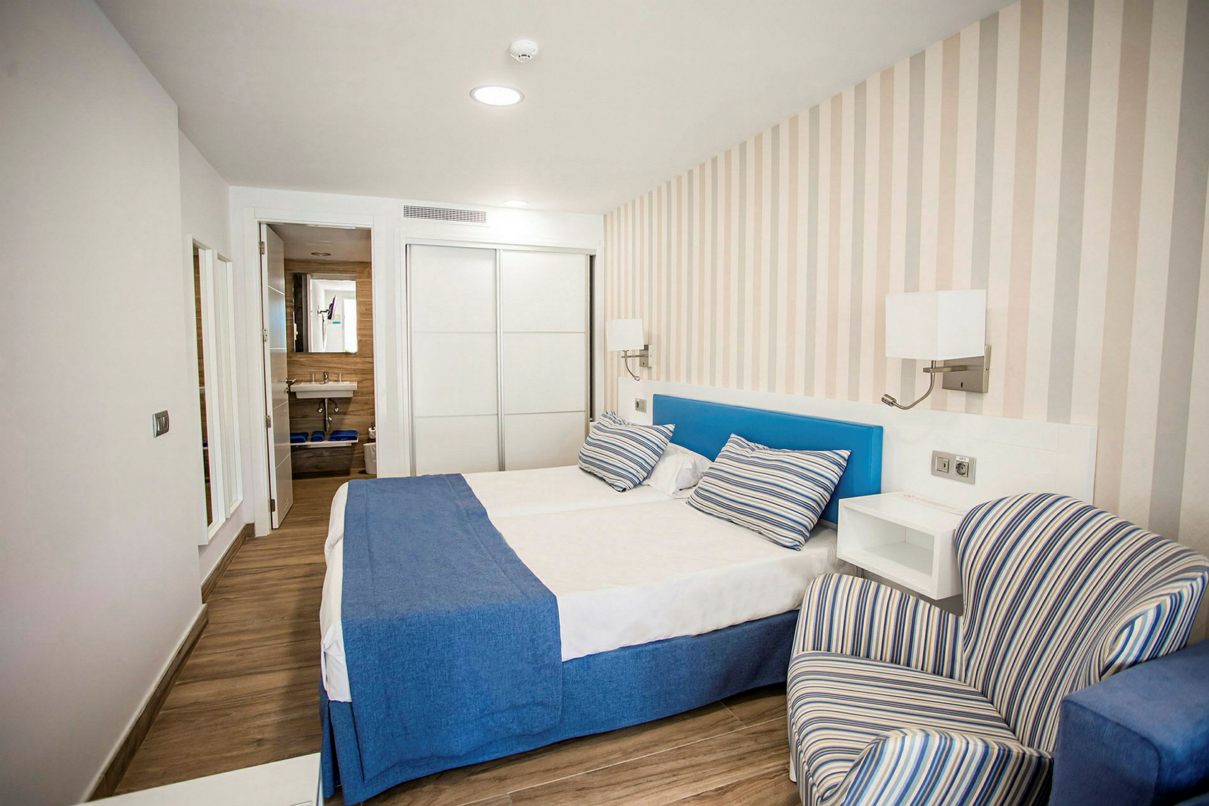 Hotellihuone, jossa on parivuode, siniset päiväpeitot ja raidalliset tyynyt, raidallinen nojatuoli sekä taustalla näkyvä kylpyhuone. (Tekoälyn luoma teksti)