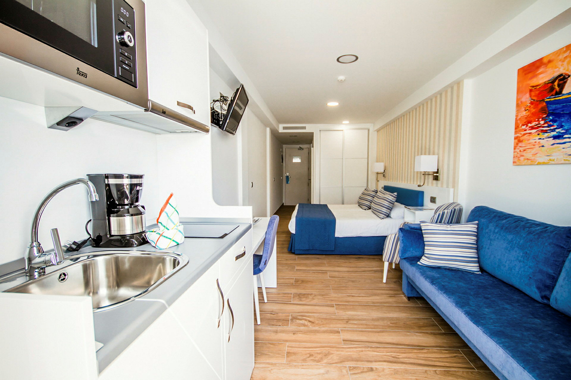 Hotellihuone, jossa on sänky, sininen sohva ja keittokomero pesualtaalla. (Tekoälyn luoma teksti)