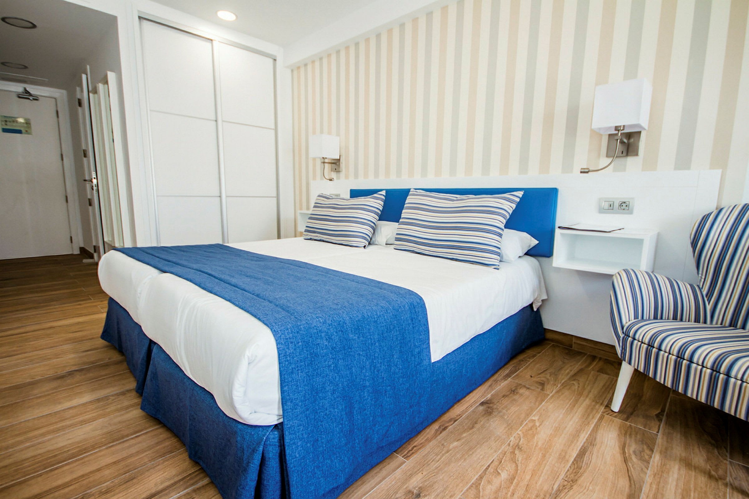 Tyylikäs hotellihuone, jossa on parivuode sinisillä ja valkoisilla vuodevaatteilla sekä raidalliset tyynyt, vieressä raidallinen nojatuoli. (Tekoälyn luoma teksti)