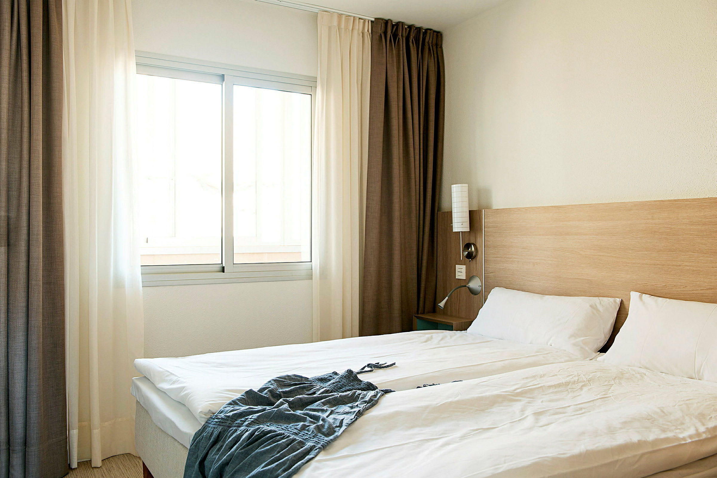 Hotellihuone, jossa on parivuode, valkoiset vuodevaatteet, puinen sängynpääty ja ikkuna vaaleilla verhoilla. (Tekoälyn luoma teksti)