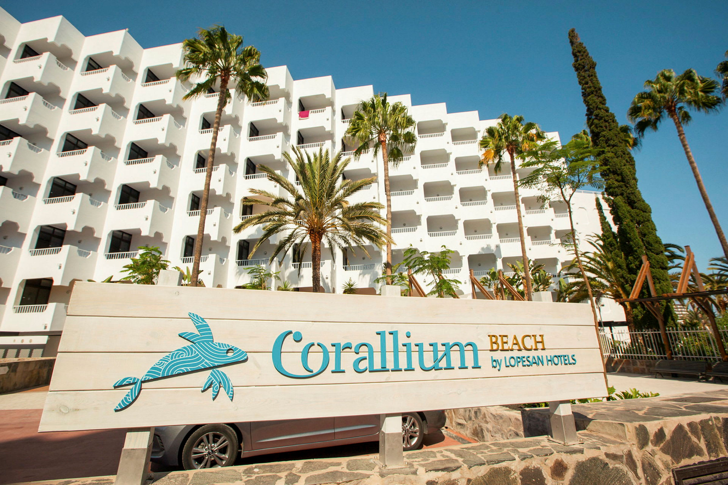 Valkoinen hotellirakennus, jossa parvekkeet, palmuja ympärillä ja kyltti, jossa lukee 'Corallium Beach by Lopesan Hotels'. (Tekoälyn luoma teksti)