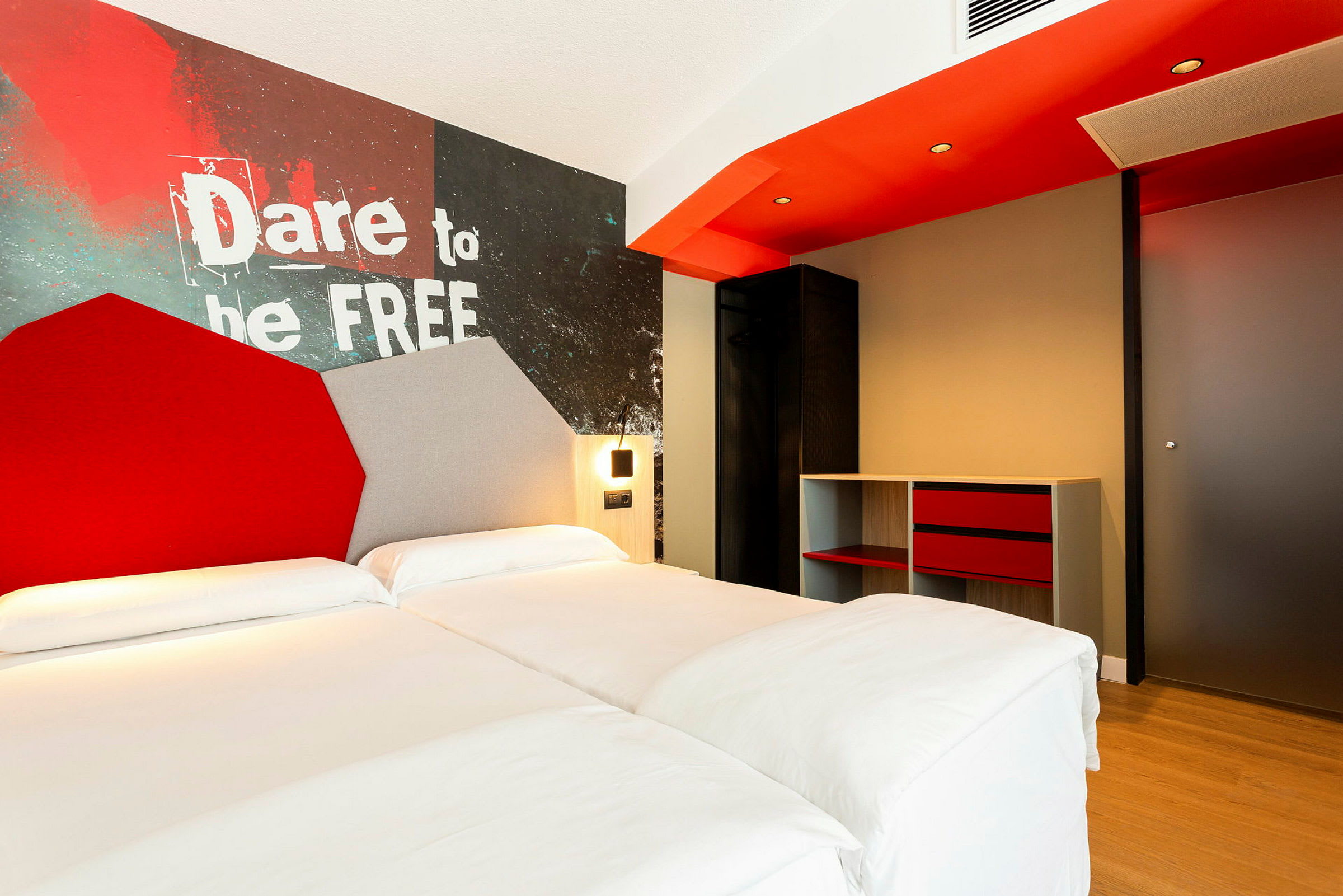 Hotellihuone, jossa on parivuode, seinäkoriste tekstillä 'Dare to be free' ja moderneja huonekaluja. (Tekoälyn luoma teksti)