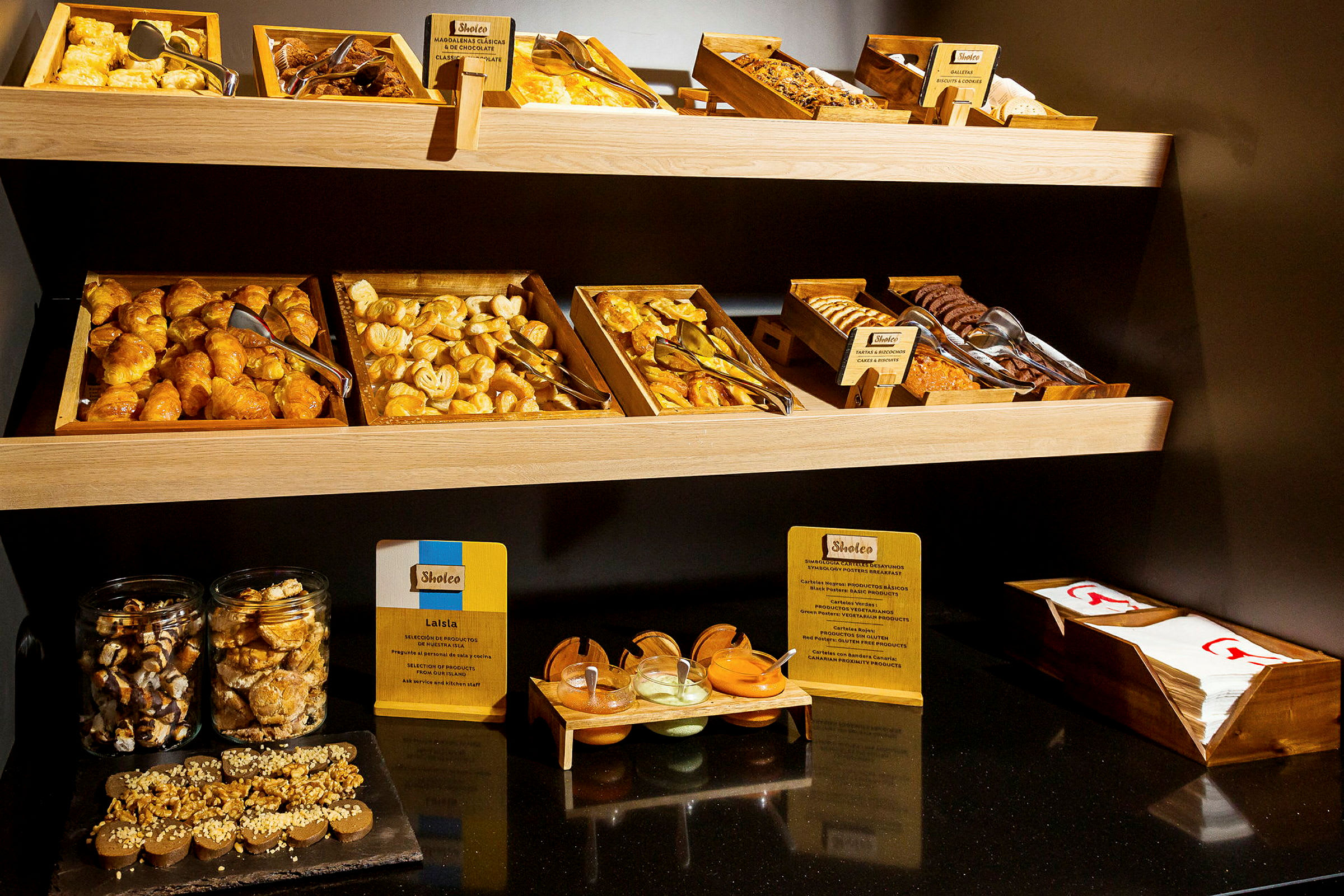 Buffet, jossa on erilaisia leivonnaisia ja lisukkeita puutasoilla, sekä purkkeja ja kulhoja erilaisilla kastikkeilla ja levitteillä. (Tekoälyn luoma teksti)