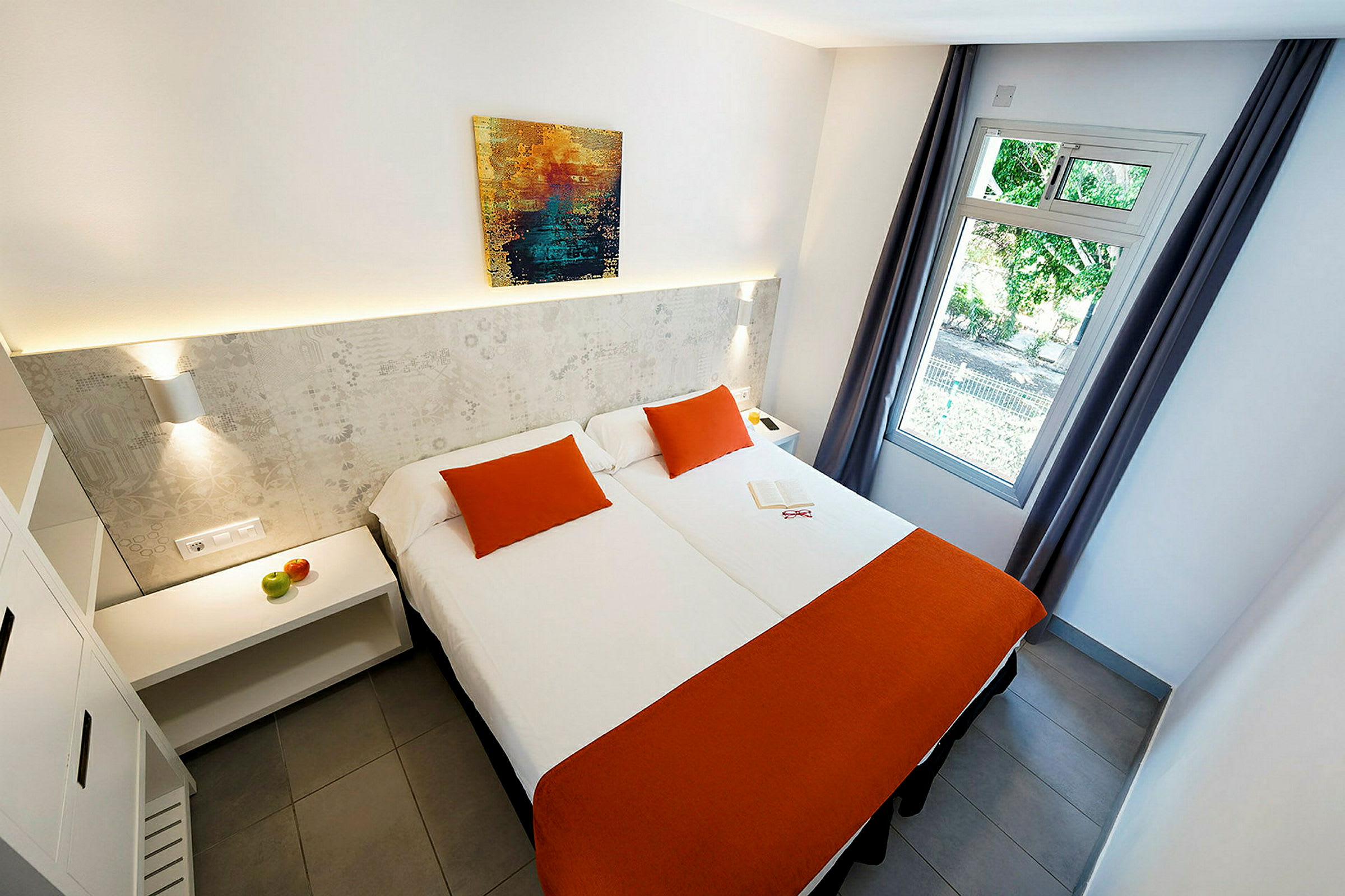 Hotellihuone, jossa kaksi sänkyä oransseilla päiväpeitteillä ja koristetyynyillä, seinällä maalaus sekä avoin ikkuna sivuverhoilla. (Tekoälyn luoma teksti)