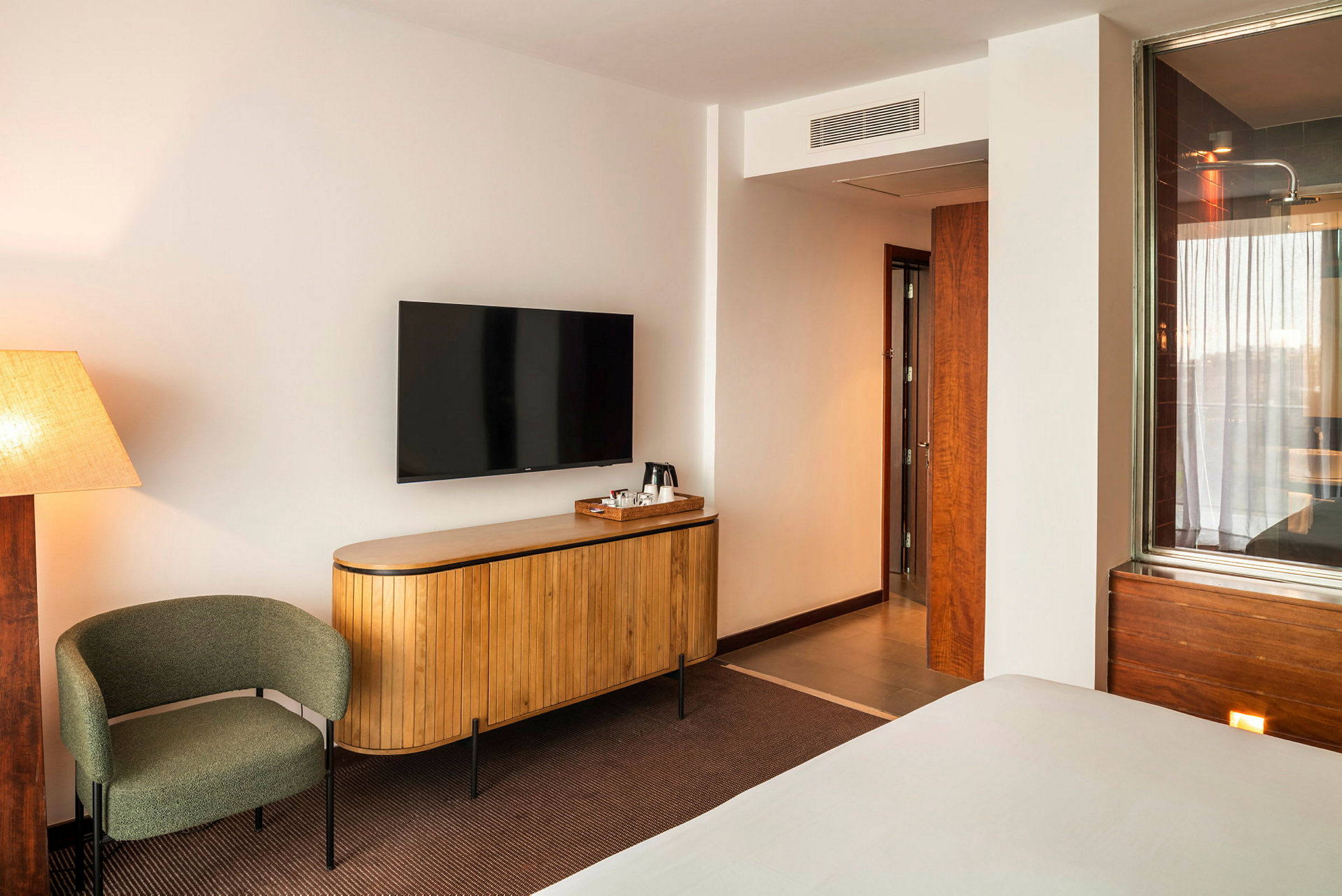 Hotellihuone, jossa on beige seinät, kääntyvä tuoli, tyylikäs seinälle asennettu televisio puisen lipaston yläpuolella ja ovi, joka johtaa kylpyhuoneeseen. (Tekoälyn luoma teksti)