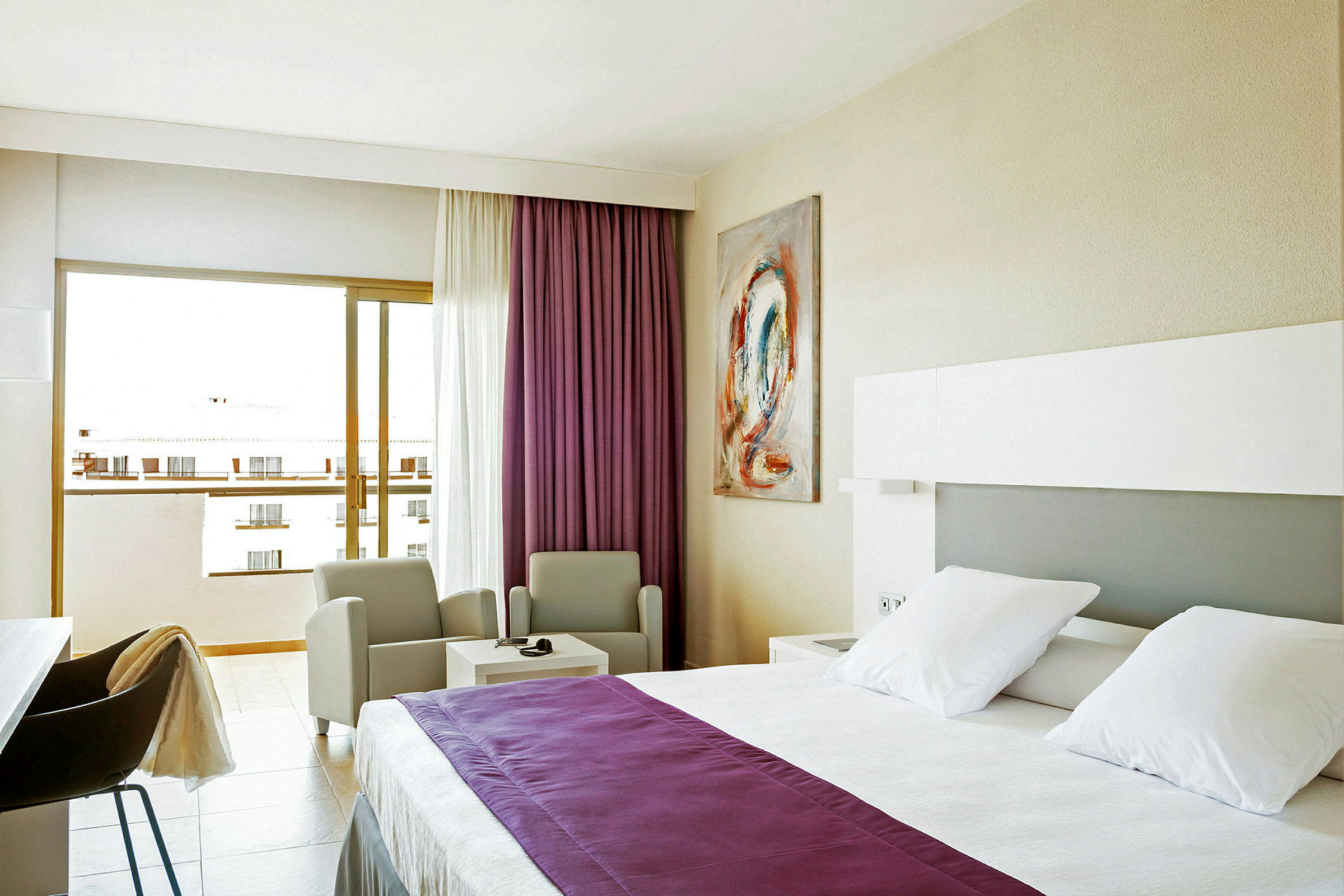 Hotellihuone, jossa on valkoiset vuodevaatteet ja violetti päiväpeite, kaksi tuolia pöydän ääressä ja parveke näkymällä. (Tekoälyn luoma teksti)