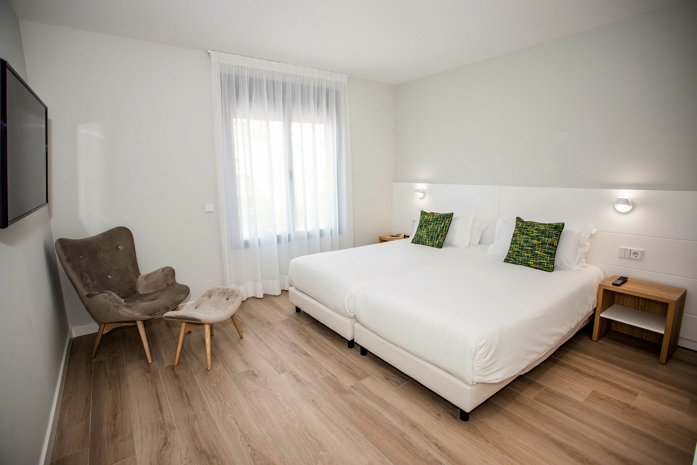 Hotellihuone, jossa kaksi yhden hengen vuodetta, nojatuoli ja jalkatuki sekä ikkuna vaaleilla verhoilla. (Tekoälyn luoma teksti)