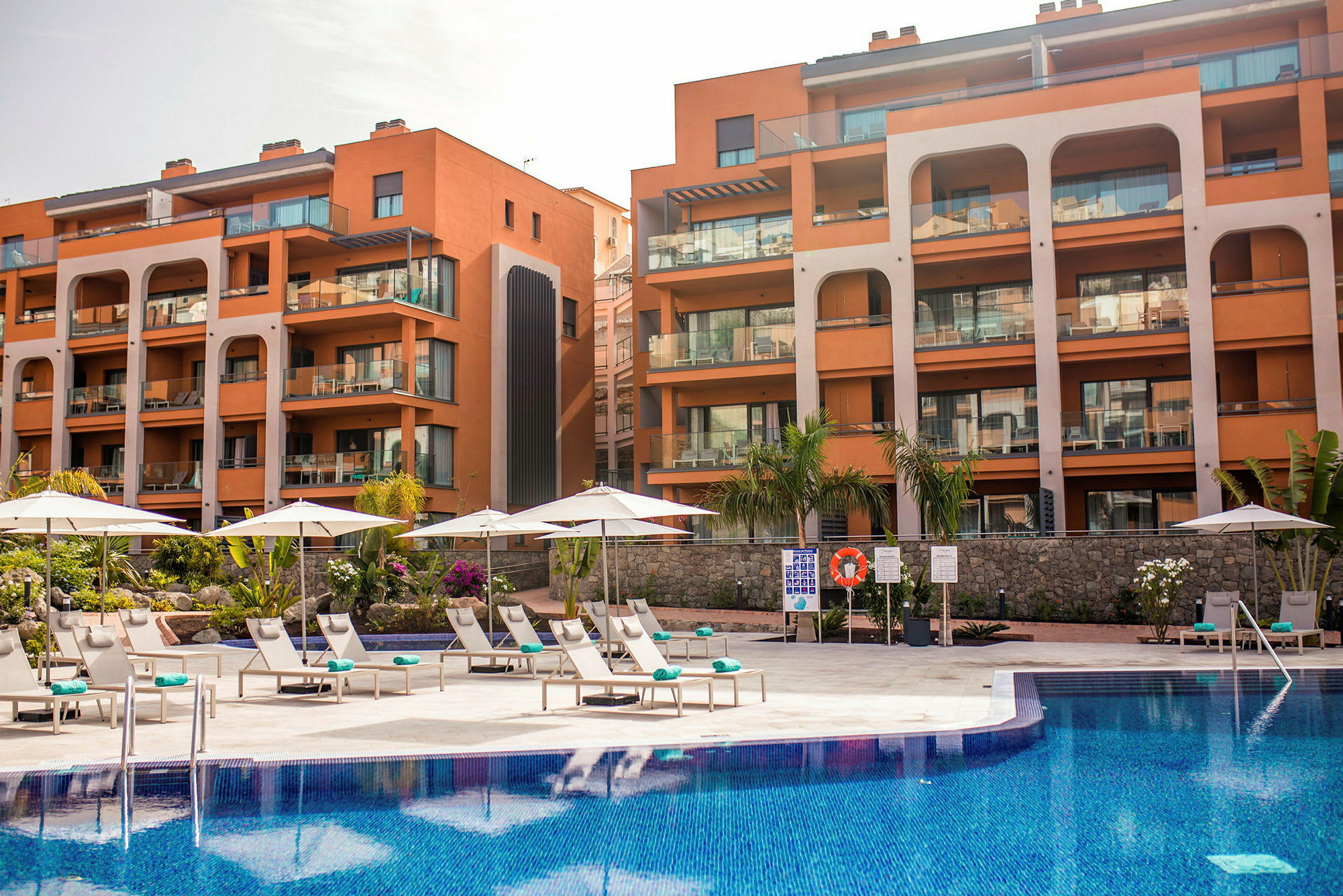 Hotellirakennus eri parvekkeilla ja edessä uima-allas, jossa aurinkotuoleja ja aurinkovarjoja. (Tekoälyn luoma teksti)