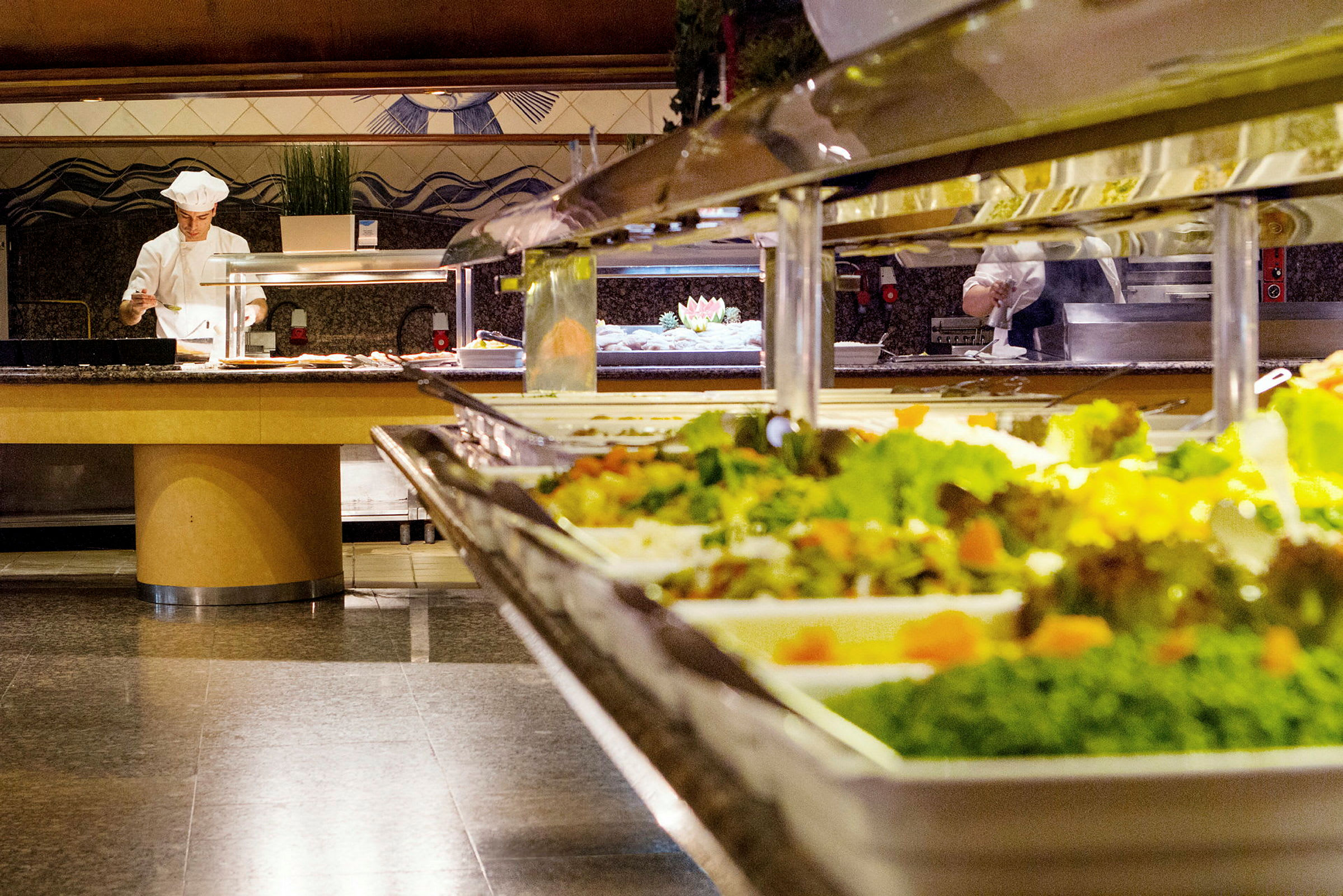 Buffet-pöytä, jossa salaatteja, ja kokki valmistamassa ruokaa taustalla. (Tekoälyn luoma teksti)