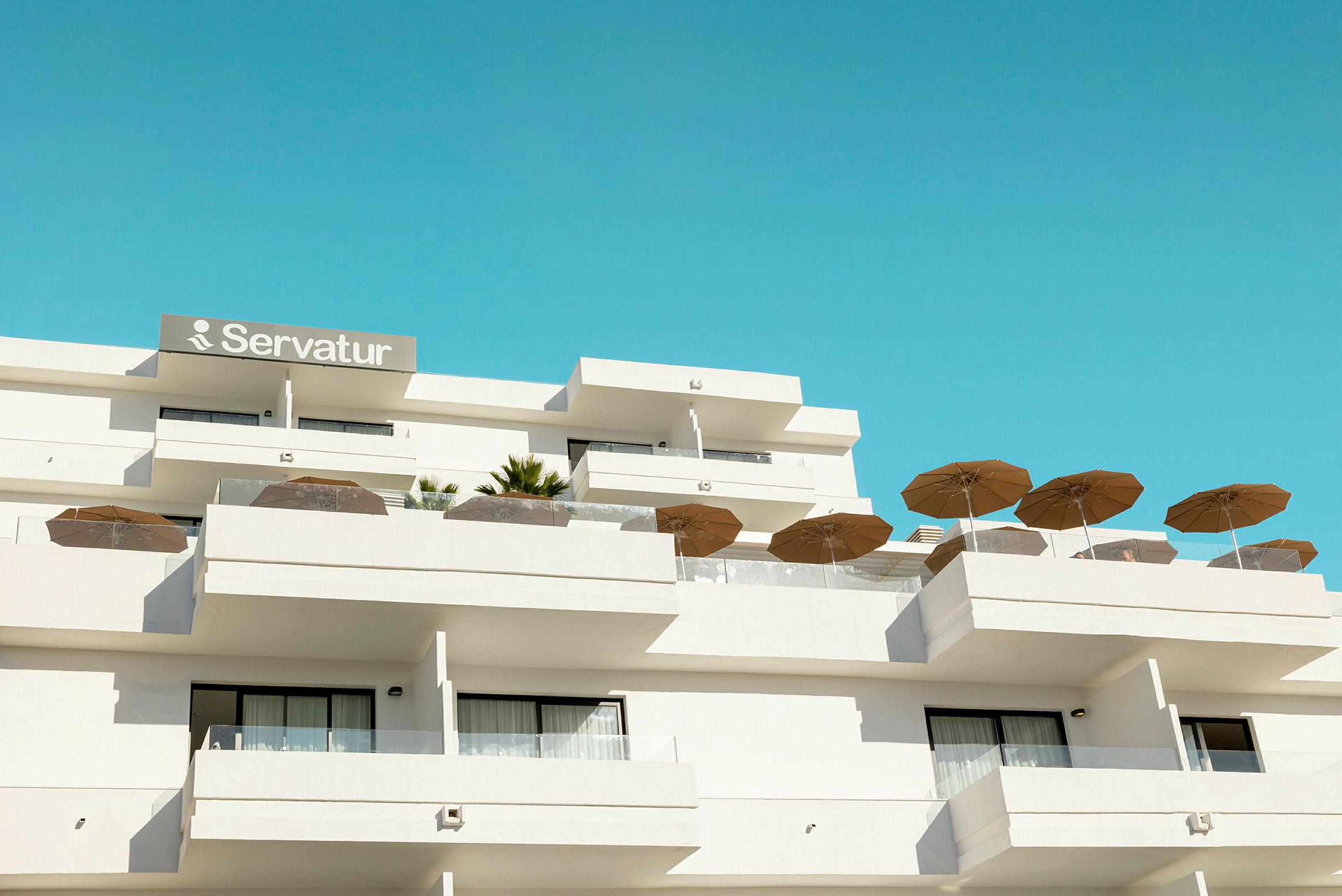 Hotellirakennus, jossa on parvekkeita ja aurinkovarjoja sinistä taivasta vasten. (Tekoälyn luoma teksti)