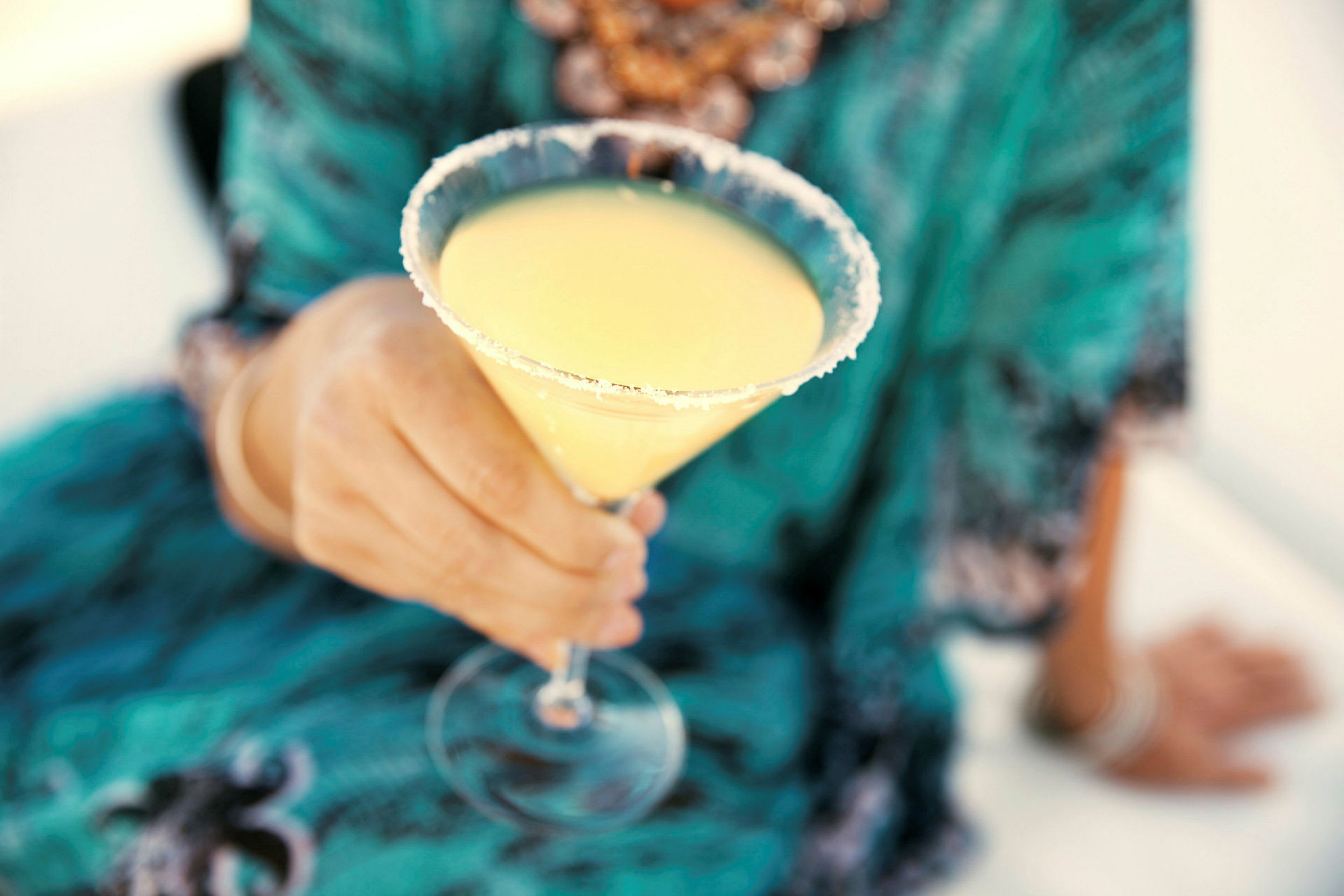 Henkilö pitää cocktail-lasista, joka on täynnä keltaista juomaa ja reunustettu sokerilla. (Tekoälyn luoma teksti)