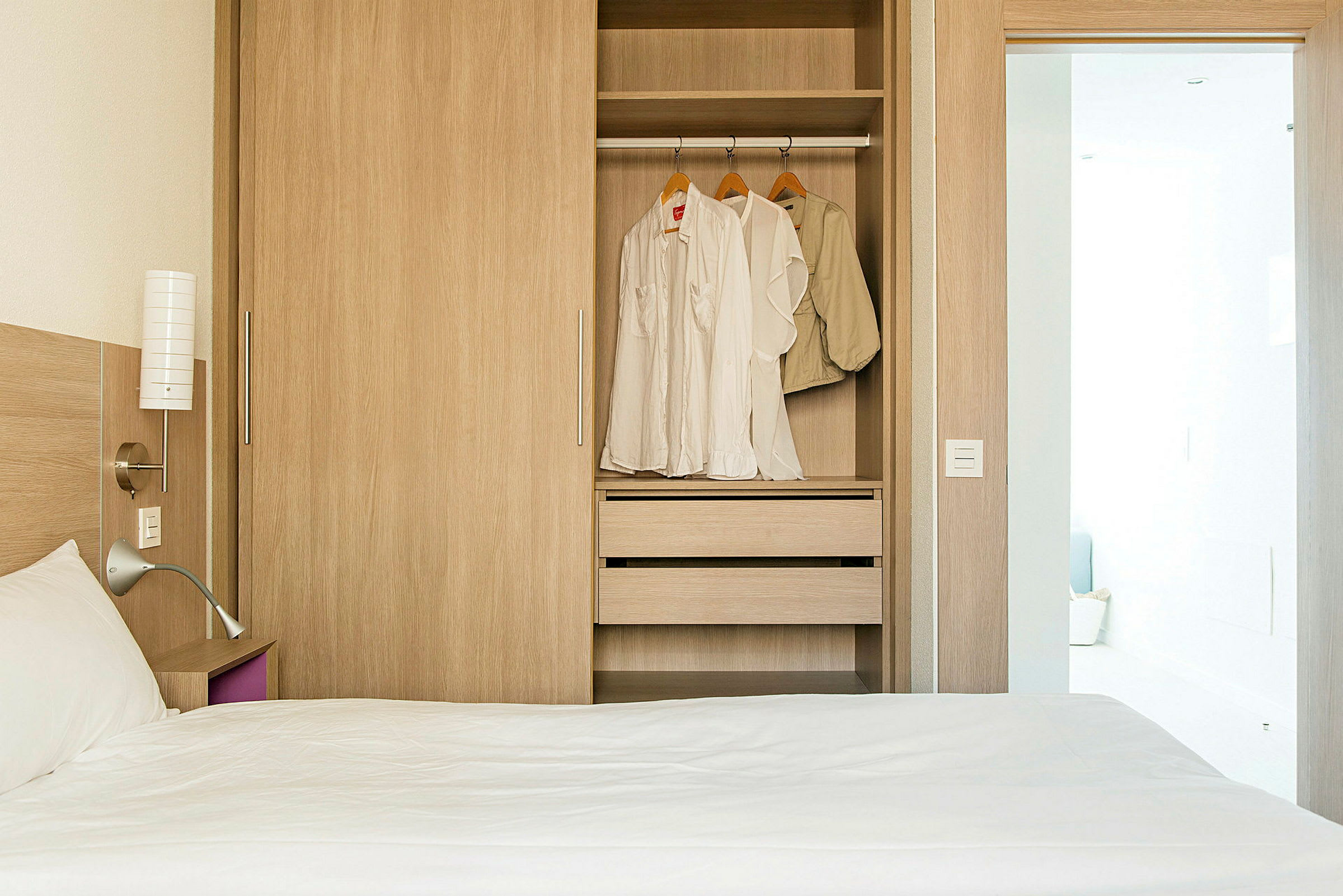 Hotellihuone, jossa on sänky ja vaatekaappi, jonka ovet ovat auki ja vaatteet roikkuvat sisällä. (Tekoälyn luoma teksti)