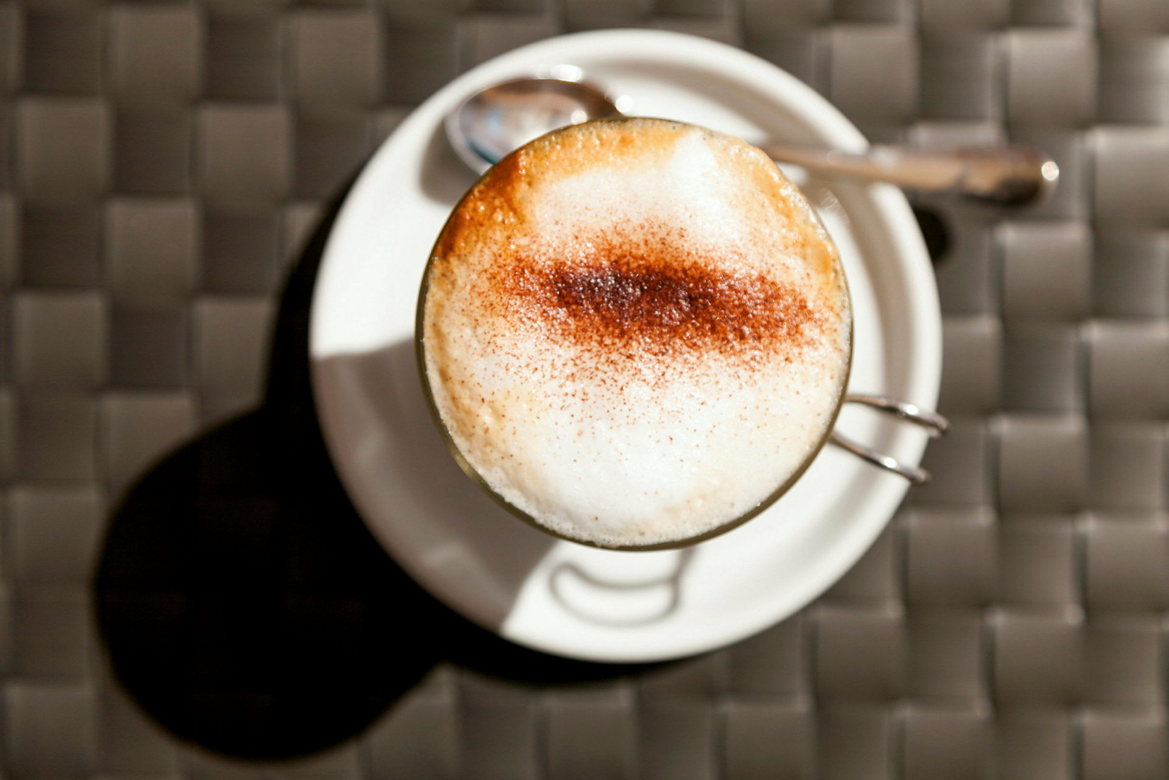 Kuppi cappuccinoa valkoisella lautasella, vieressä lusikka. (Tekoälyn luoma teksti)