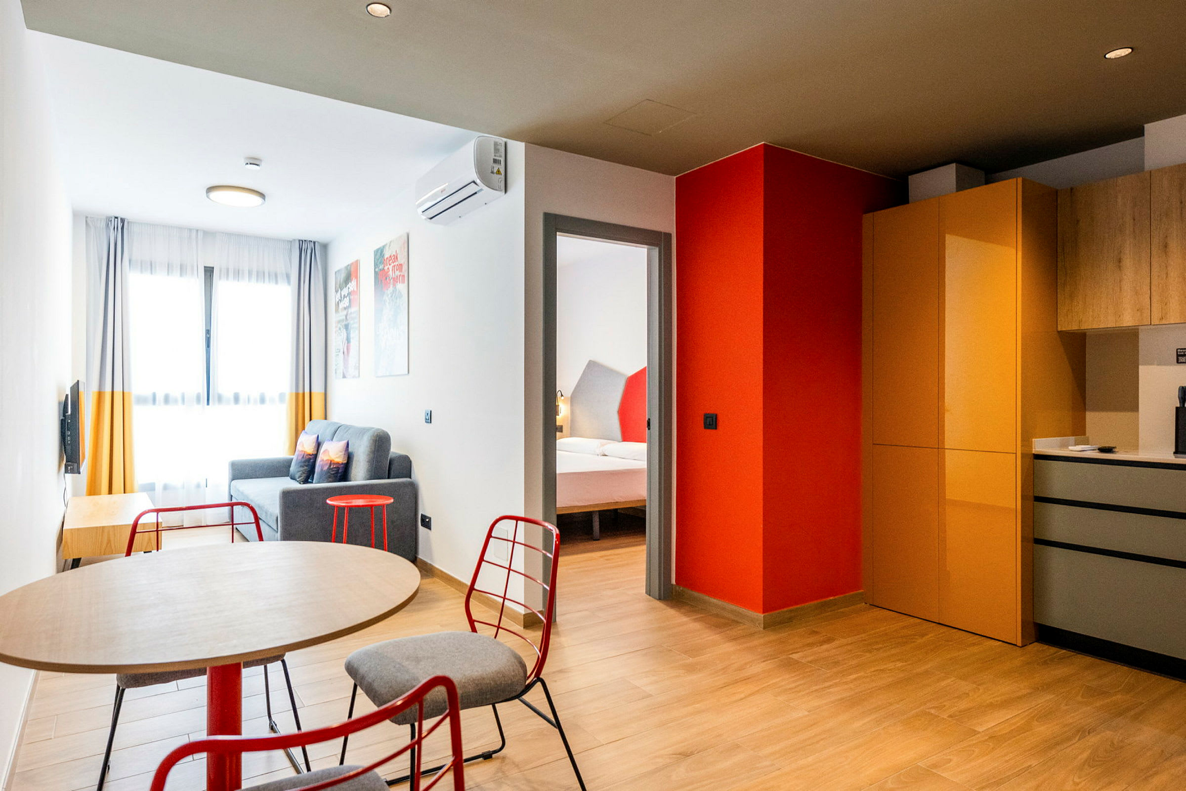 Modernisti sisustettu hotellihuone, jossa etualalla pöytä ja tuolit, istuinryhmä sohvineen ja makuuhuone näkyy oviaukosta. Keittiöalueessa on kaapit ja työtilat. (Tekoälyn luoma teksti)