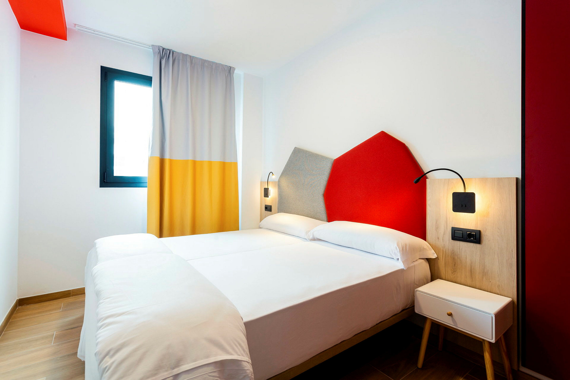 Hotellihuone, jossa on parivuode, värikkäitä seinäkoristeita ja ikkuna verhoineen. (Tekoälyn luoma teksti)