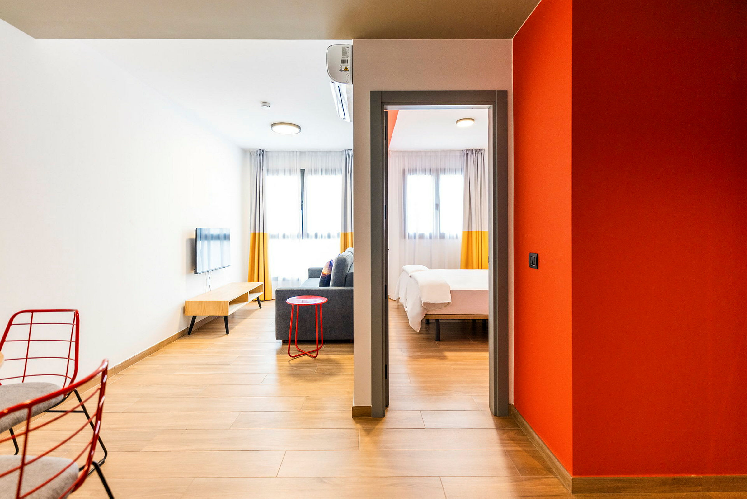 Moderni hotellihuone, jossa on puulattia ja viihtyisä sisustus. Huoneessa on oleskelualue sohvalla, seinälle kiinnitetty televisio ja makuuhuone, jossa on parisänky. (Tekoälyn luoma teksti)