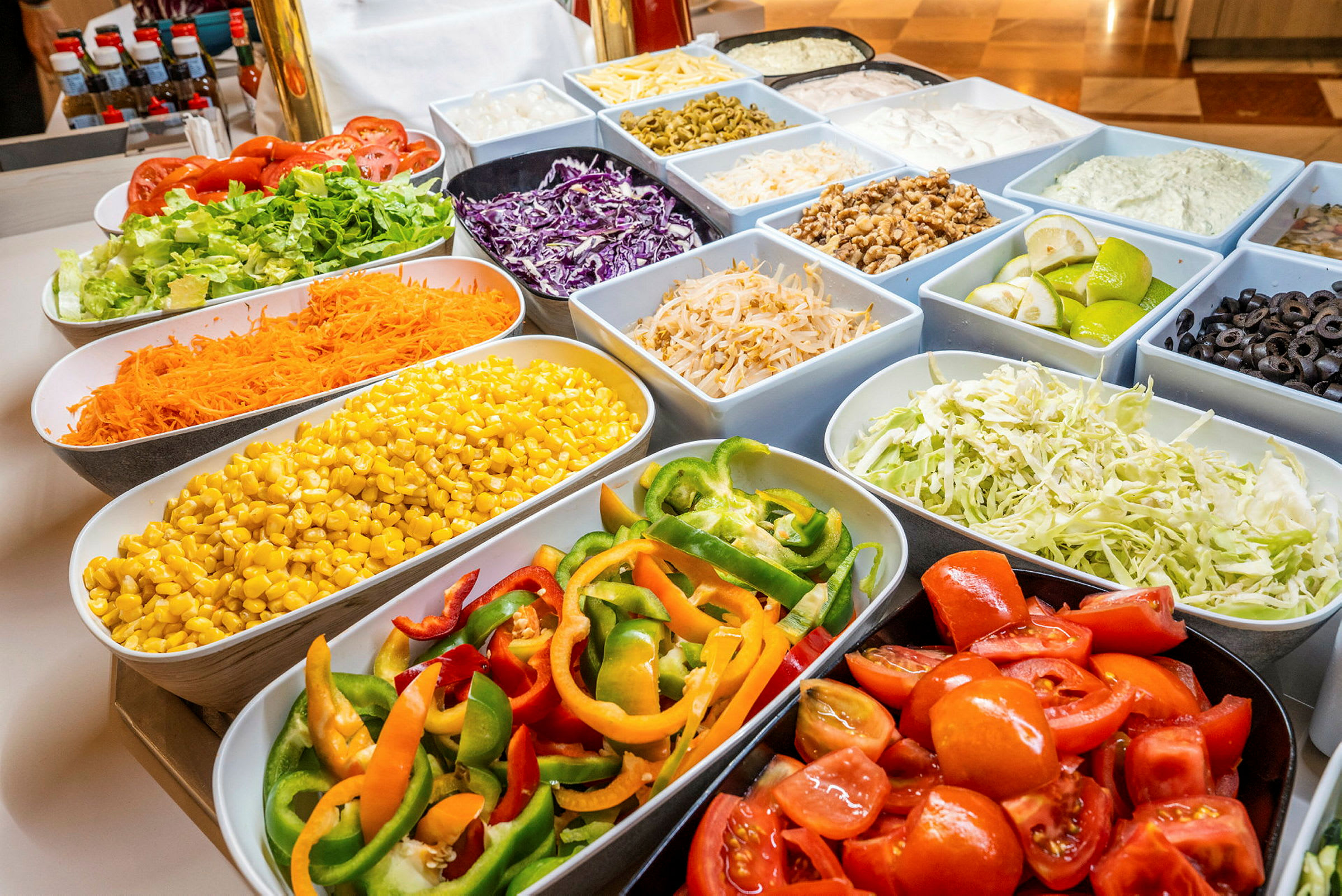 Buffet-pöytä, jossa on erilaisia vihanneksia kulhoissa, mukaan lukien tomaatit, salaatti, maissi, paprika ja oliivit. (Tekoälyn luoma teksti)