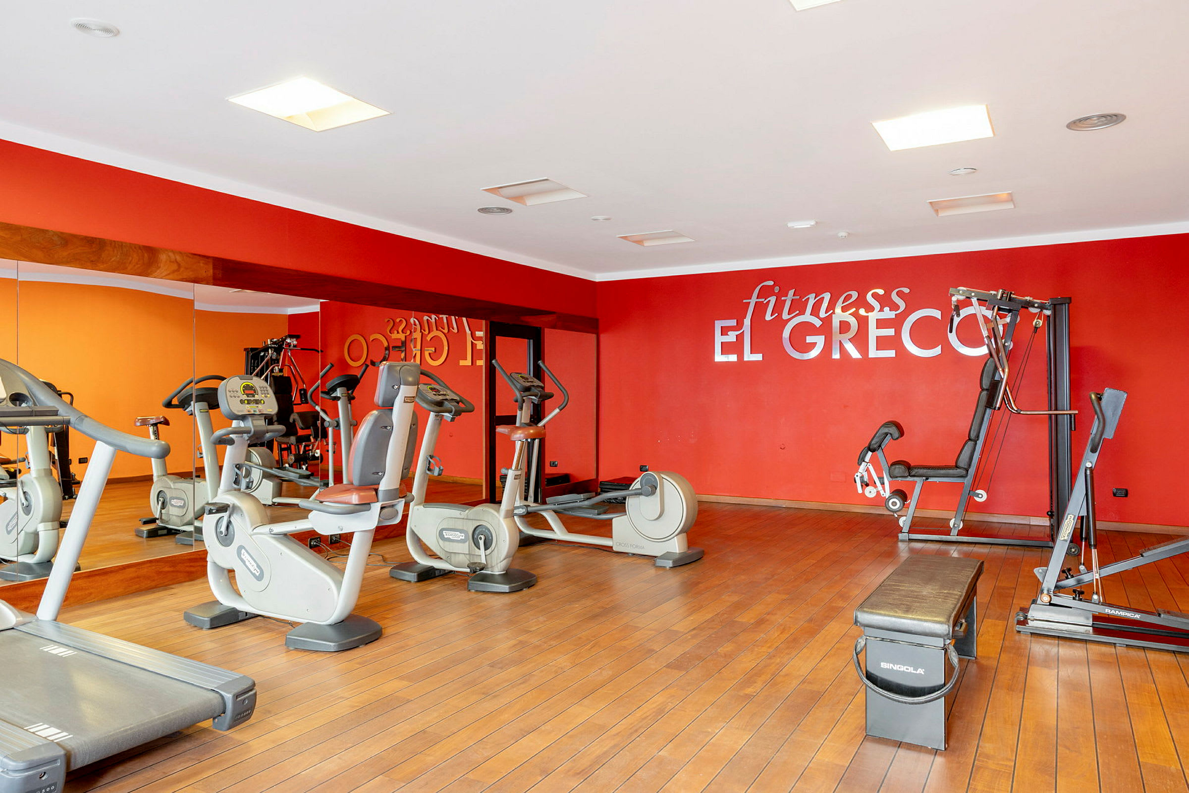 Kuntosali, jossa kuntosalilaitteita, peilejä ja punainen seinä tekstillä 'fitness El Greco'. (Tekoälyn luoma teksti)
