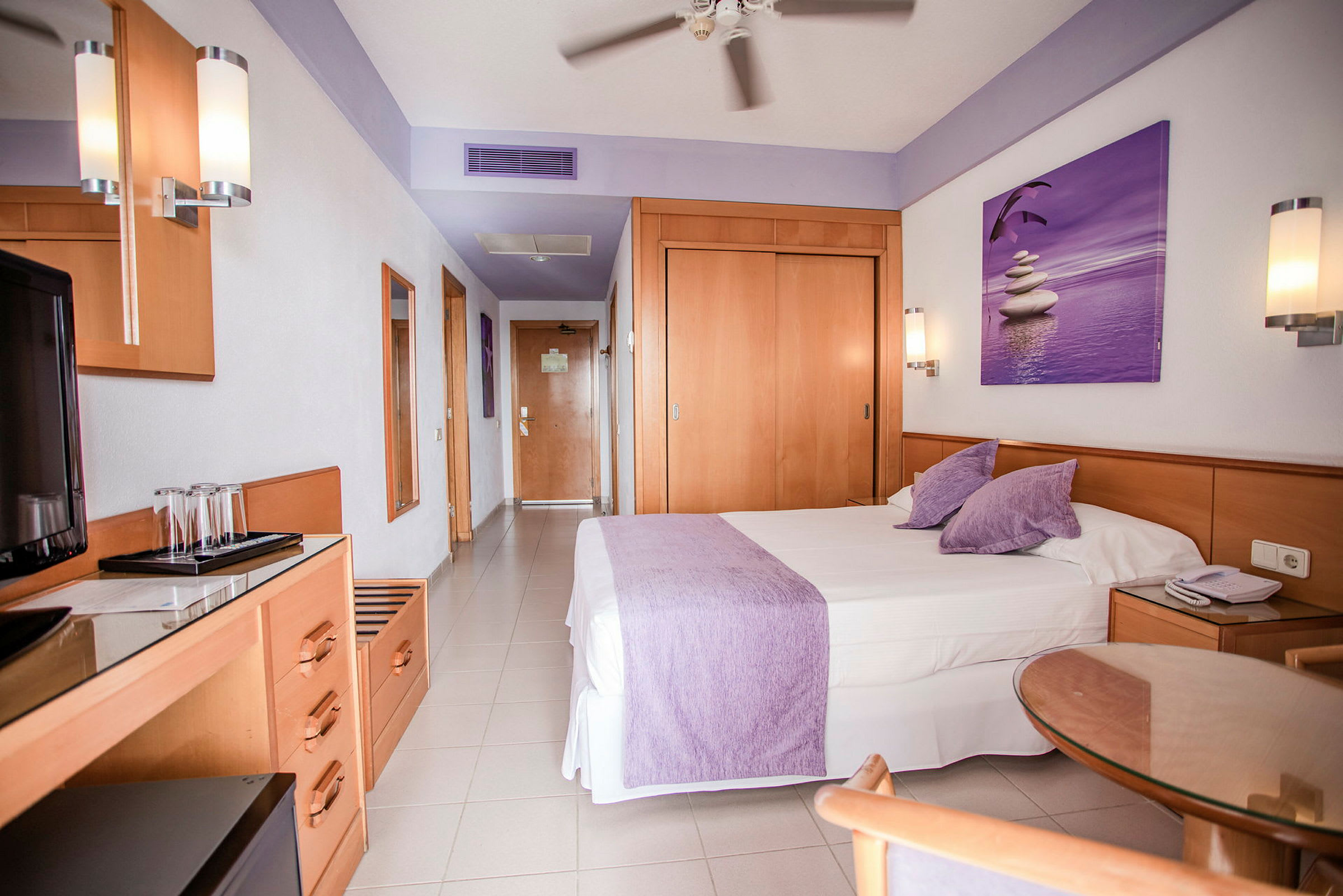Hotellihuone, jossa on suuri sänky violeteilla vuodevaatteilla, puuhuonekaluja ja koristeita. (Tekoälyn luoma teksti)