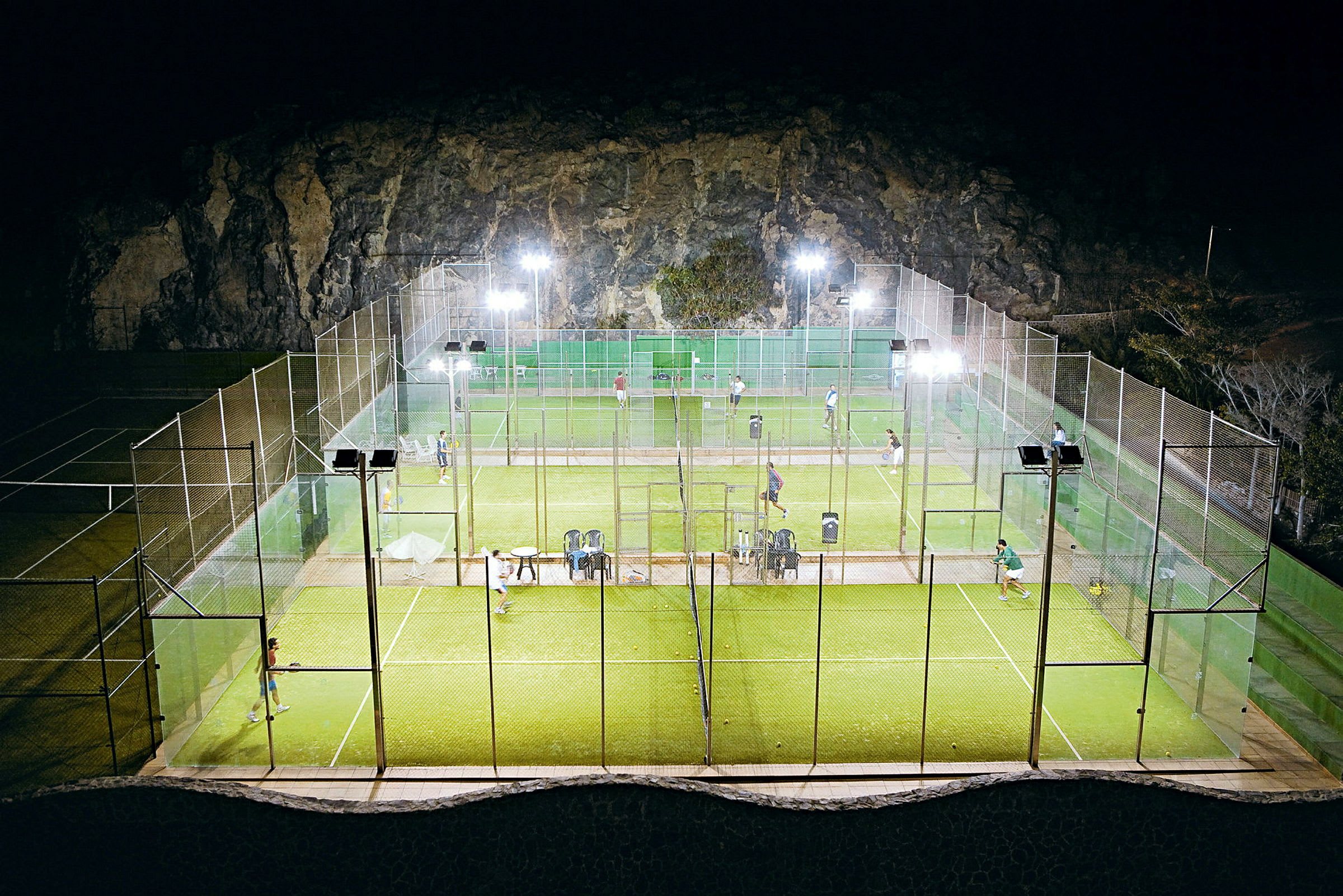 Ulkopadelkentät valaistuna illalla, pelaajia näkyvissä kentällä. (Tekoälyn luoma teksti)