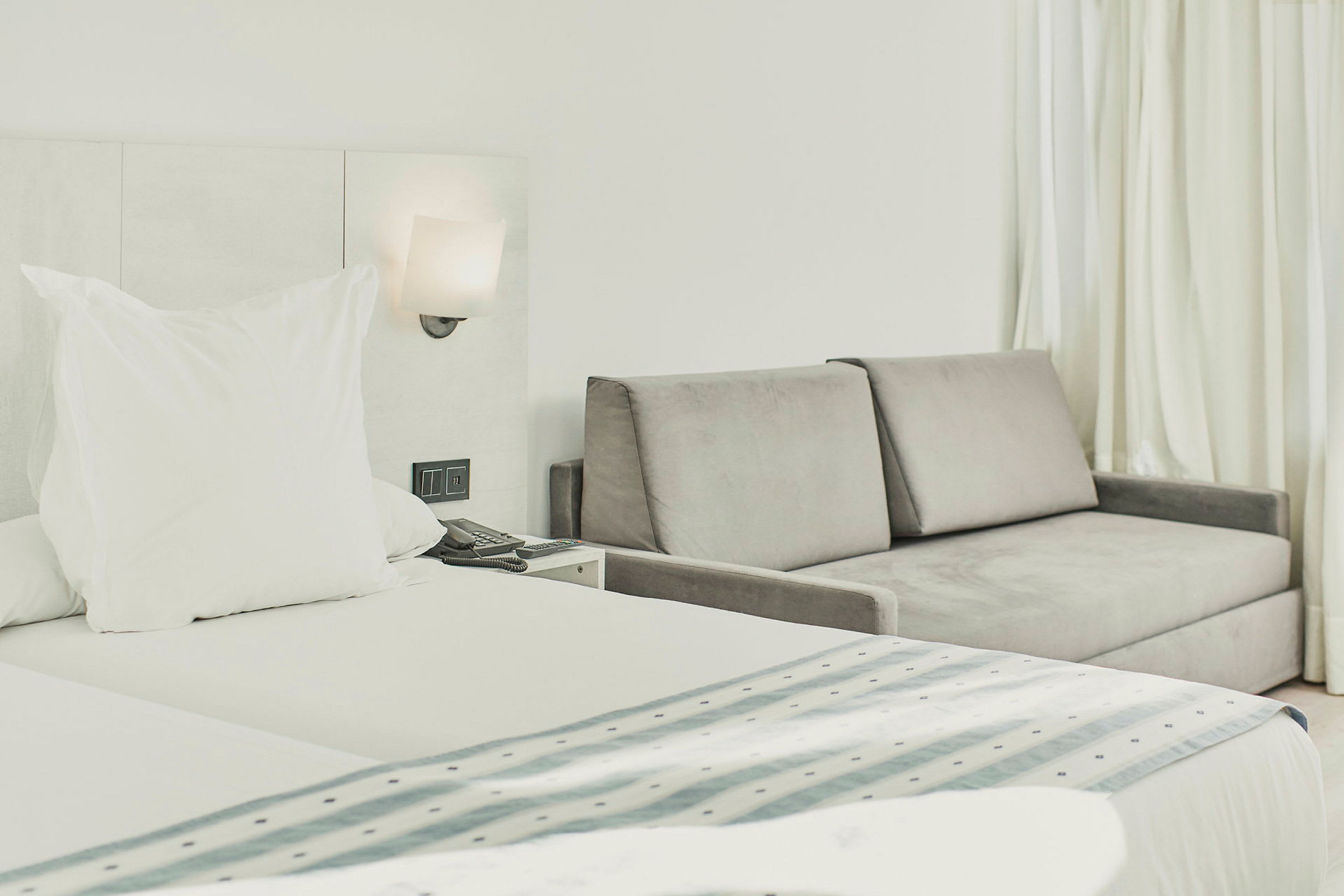 Hotellihuone, jossa sänky ja harmaa sohva vieressä. (Tekoälyn luoma teksti)