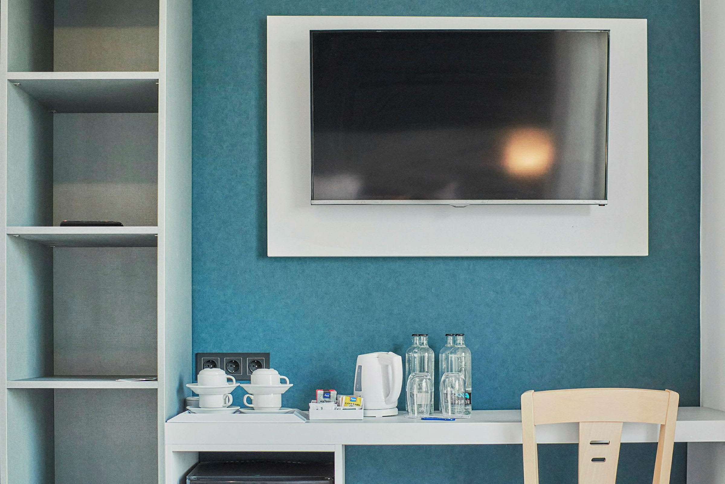 Hotellihuone, jossa seinälle asennettu TV, valkoinen huonekalu, jossa vedenkeitin, kupit ja pullot, ja sivussa avoin kaappi. (Tekoälyn luoma teksti)