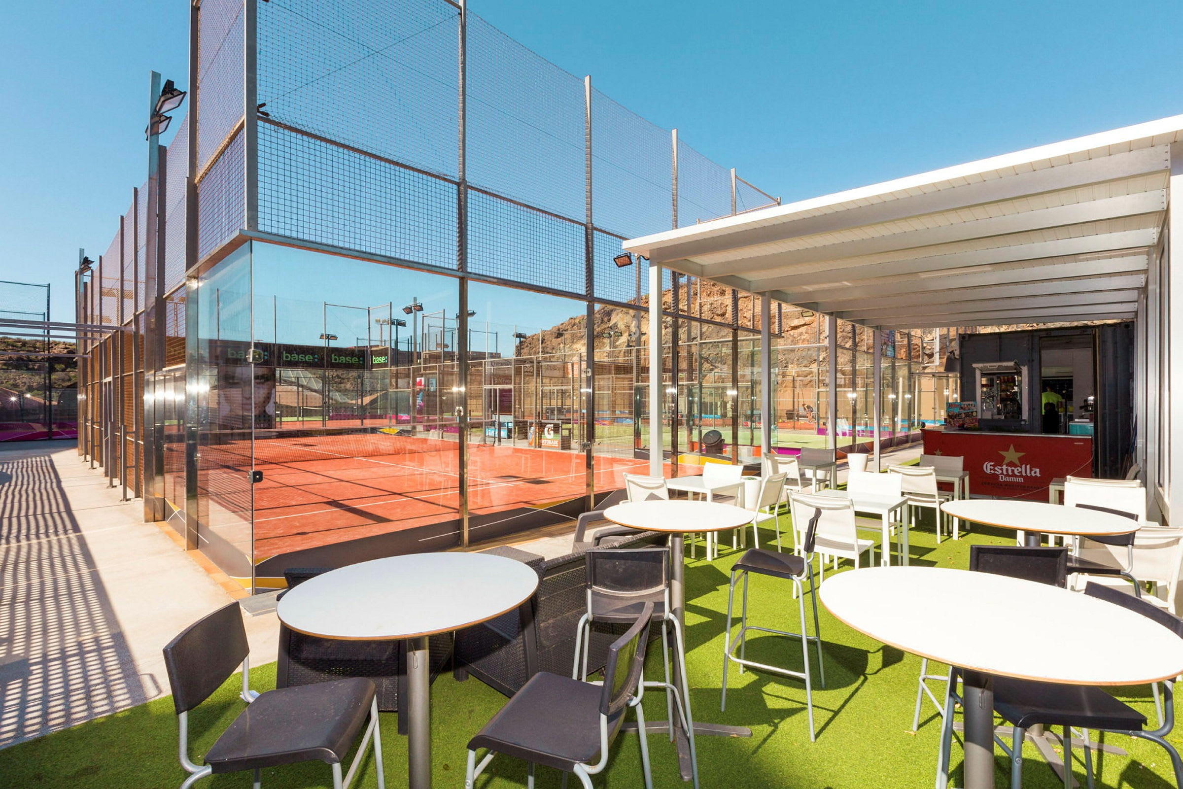 Ulkoilmaravintola padelkentän vieressä, jossa vihreä tekonurmimatto ja valkoiset kalusteet katoksen alla. (Tekoälyn luoma teksti)