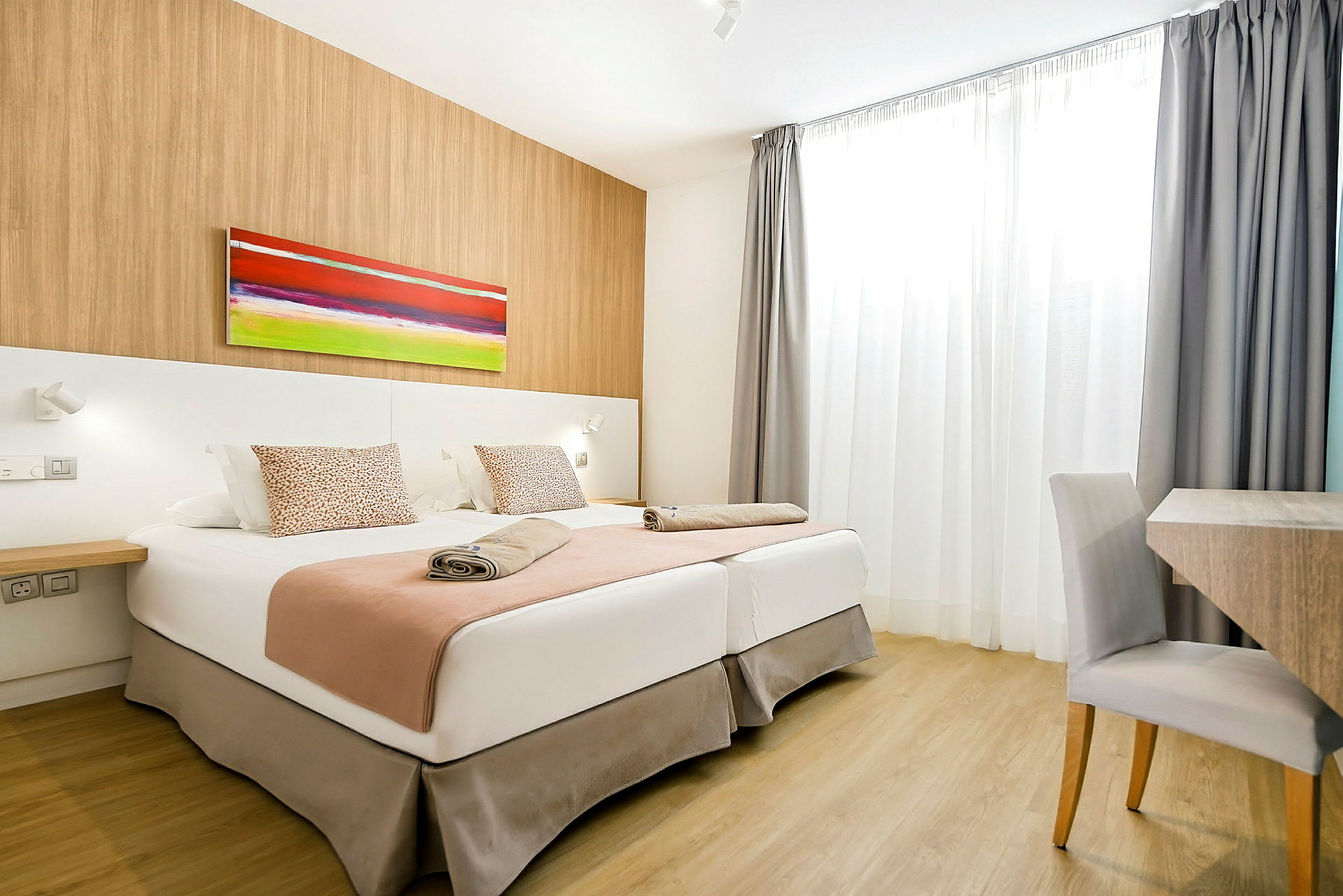 Hotellihuone, jossa kaksi vuodetta, värikäs taulu seinällä ja kirjoituspöytä tuolilla. (Tekoälyn luoma teksti)