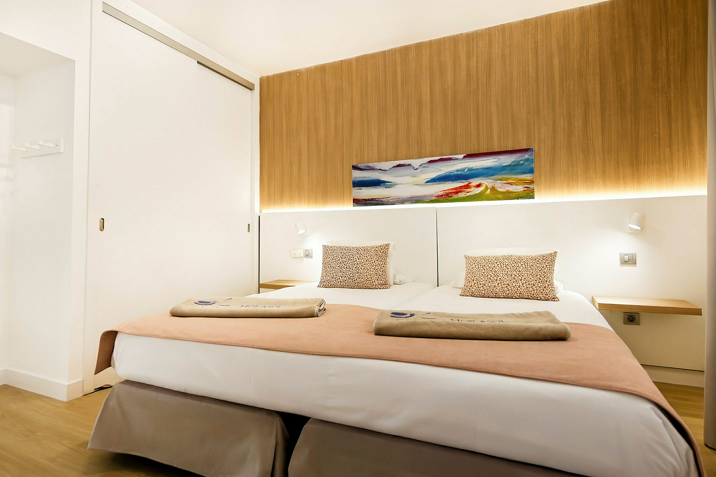 Moderni hotellihuone, jossa on kaksi erillistä sänkyä, koristeltuina tyynyillä ja pyyhkeillä, taulu seinällä ja taustalla puupaneelia. (Tekoälyn luoma teksti)
