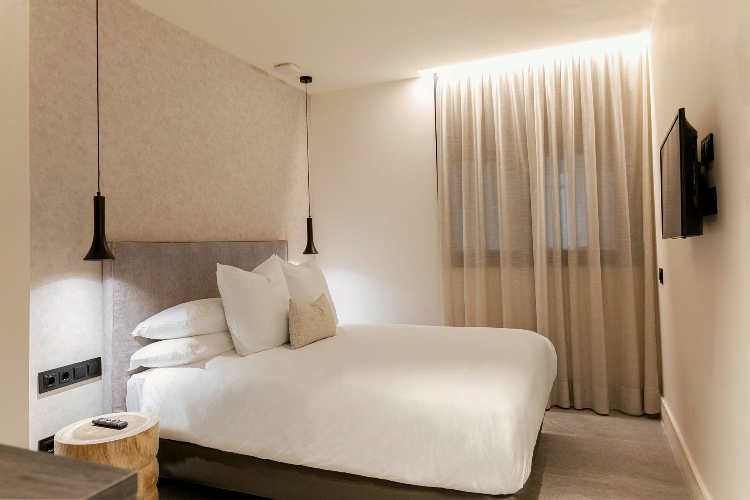 Hotellihuone, jossa on parisänky, kaksi roikkuvaa lamppua, ikkuna läpinäkyvien verhojen kanssa ja seinään kiinnitetty televisio. (Tekoälyn luoma teksti)