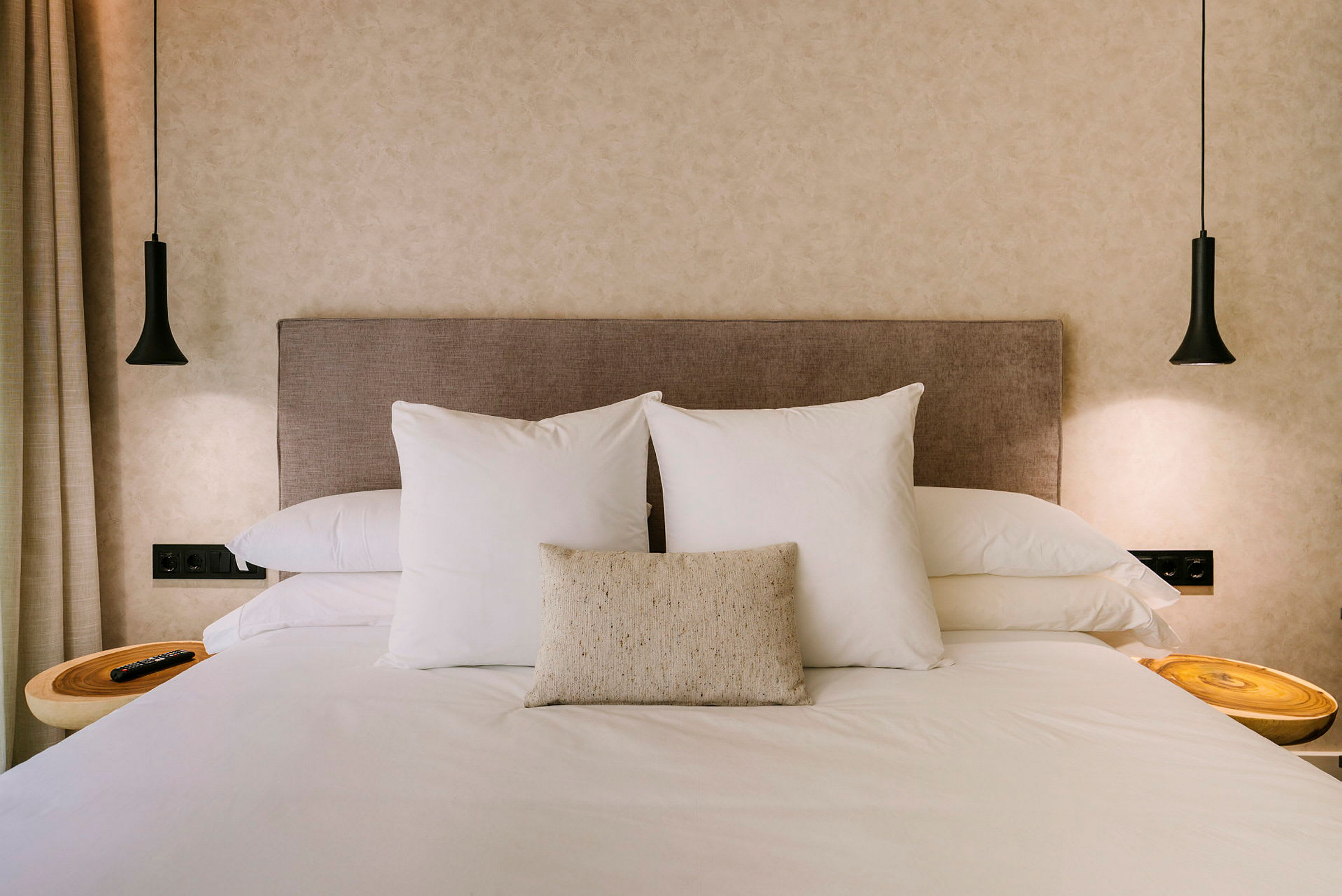 Hotellisänky, jossa kaksi suurta valkoista tyynyä ja yksi pienempi koristetyyny beigellä päätylevyllä. (Tekoälyn luoma teksti)