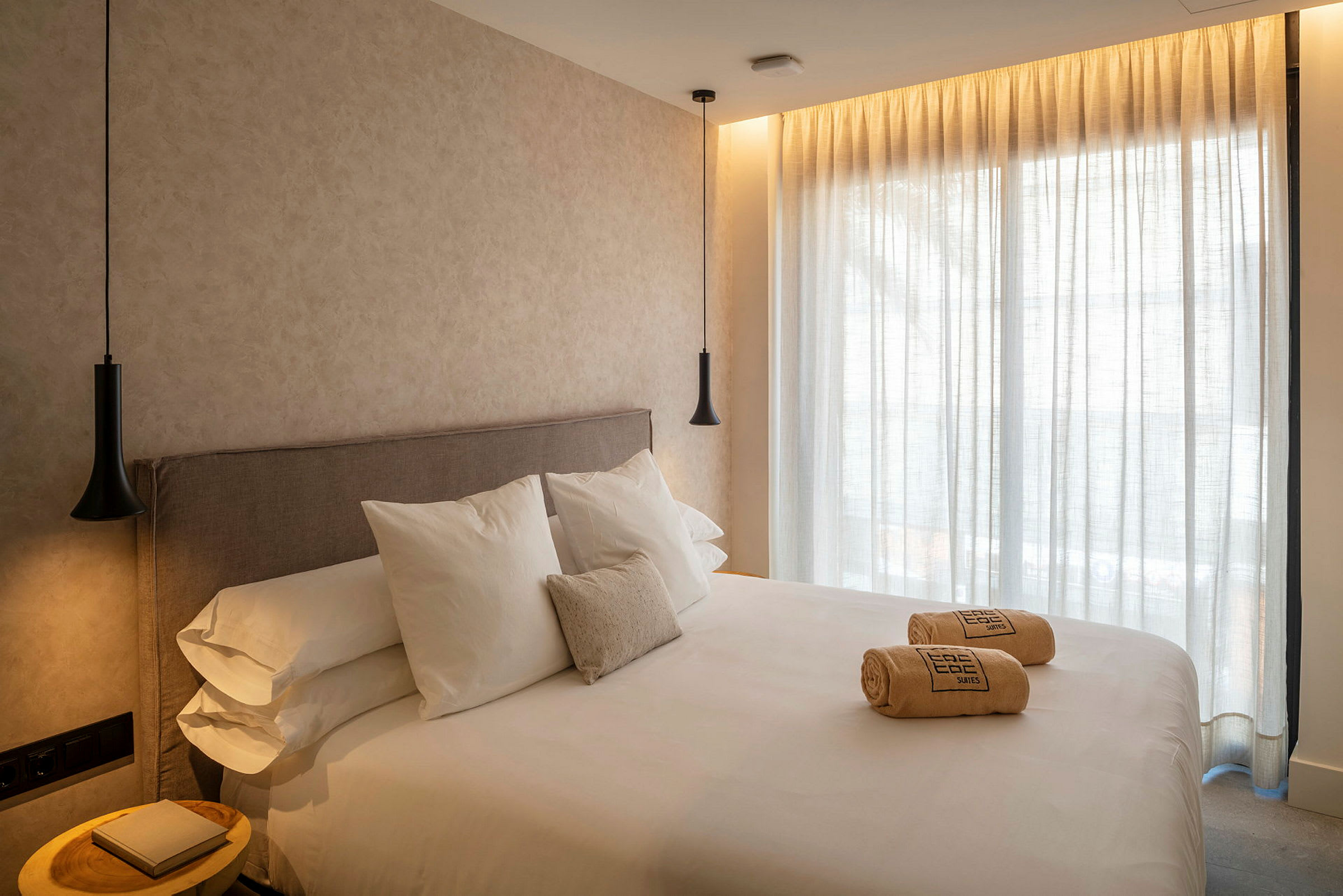 Hotellihuone, jossa on suuri sänky, valkoiset lakanat ja tyynyt. Sängyllä on rullattuja pyyhkeitä. Seinät ja verhot ovat vaaleita ja huoneessa on riippuvalaisimet. (Tekoälyn luoma teksti)