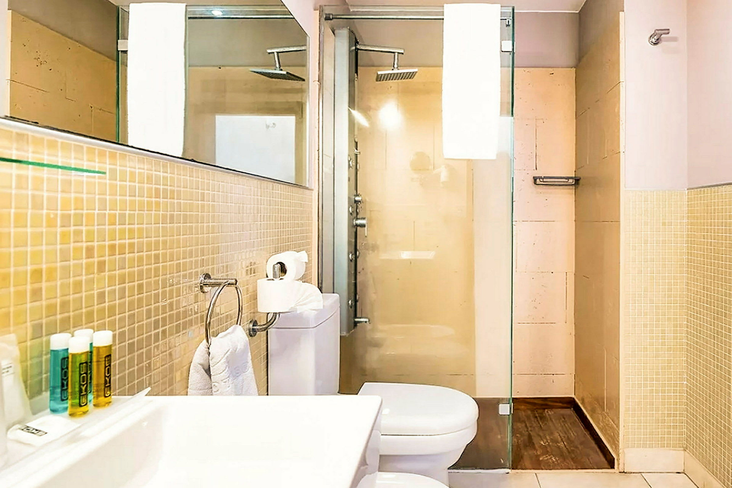 Kuva kylpyhuoneesta, jossa on suihku, wc ja pesuallas. Beige seinät ja lasiovinen suihkukaappi. (Tekoälyn luoma teksti)